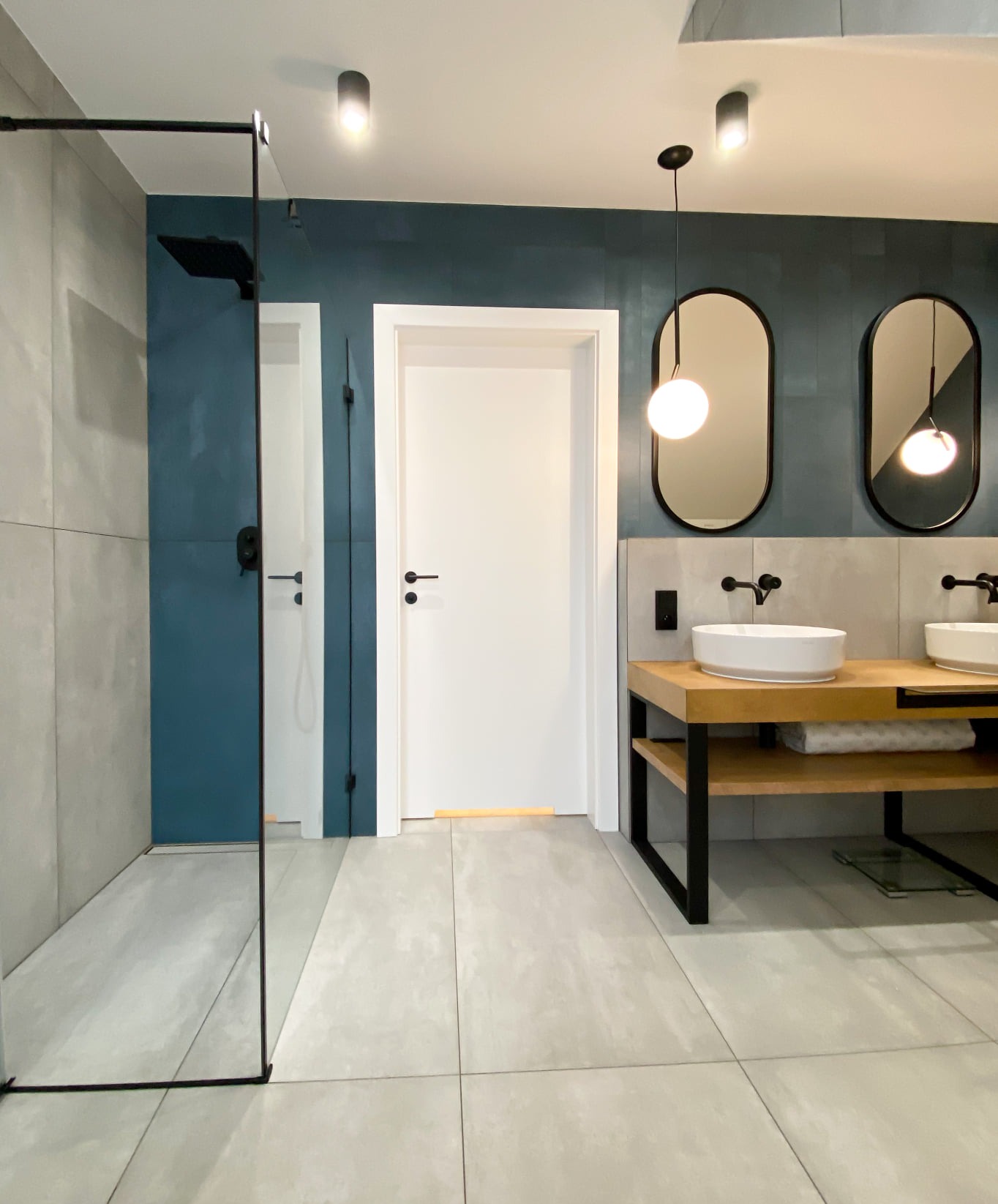 Nowoczesna łazienka z kabiną prysznicową typu walk-in, podwójną umywalką na drewnianym blacie z czarnymi nogami, owalnymi lustrami podwieszonymi na czarnych przewodach oraz niebiesko-szarą ścianą.
