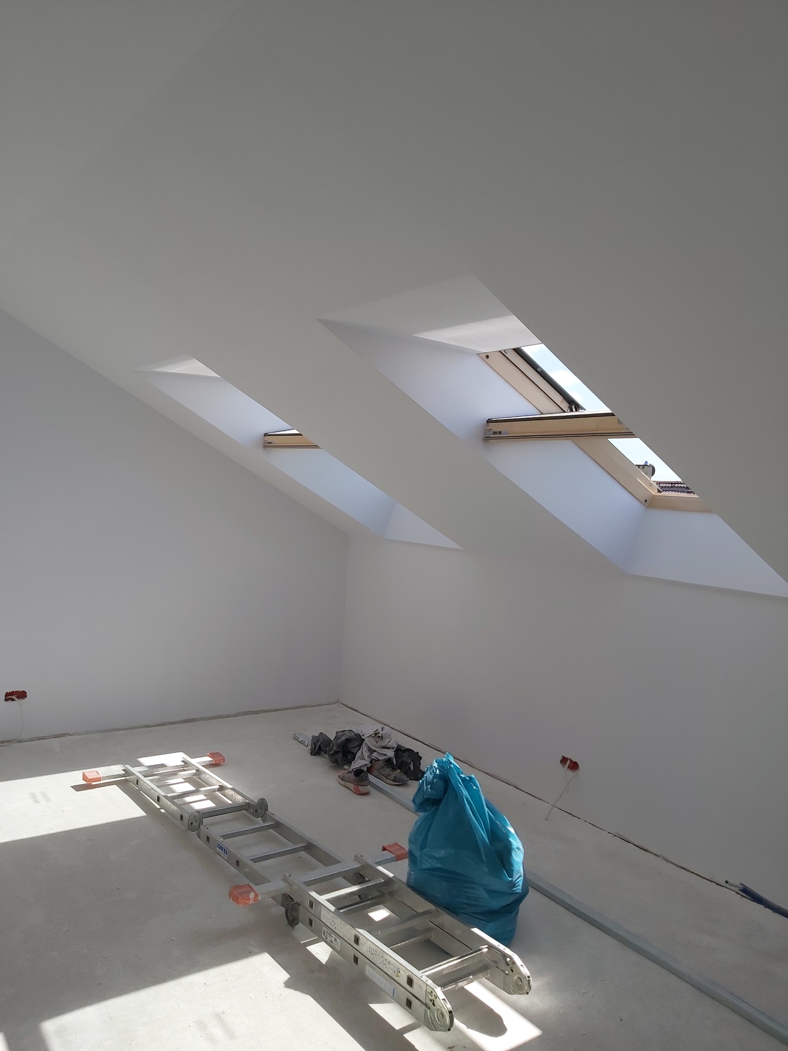 Poddasze po świeżym pomalowaniu na biało, widoczne dwa okna dachowe, rozłożona drabina aluminiowa, niebieski worek na śmieci i ślady po gładzi na podłodze.