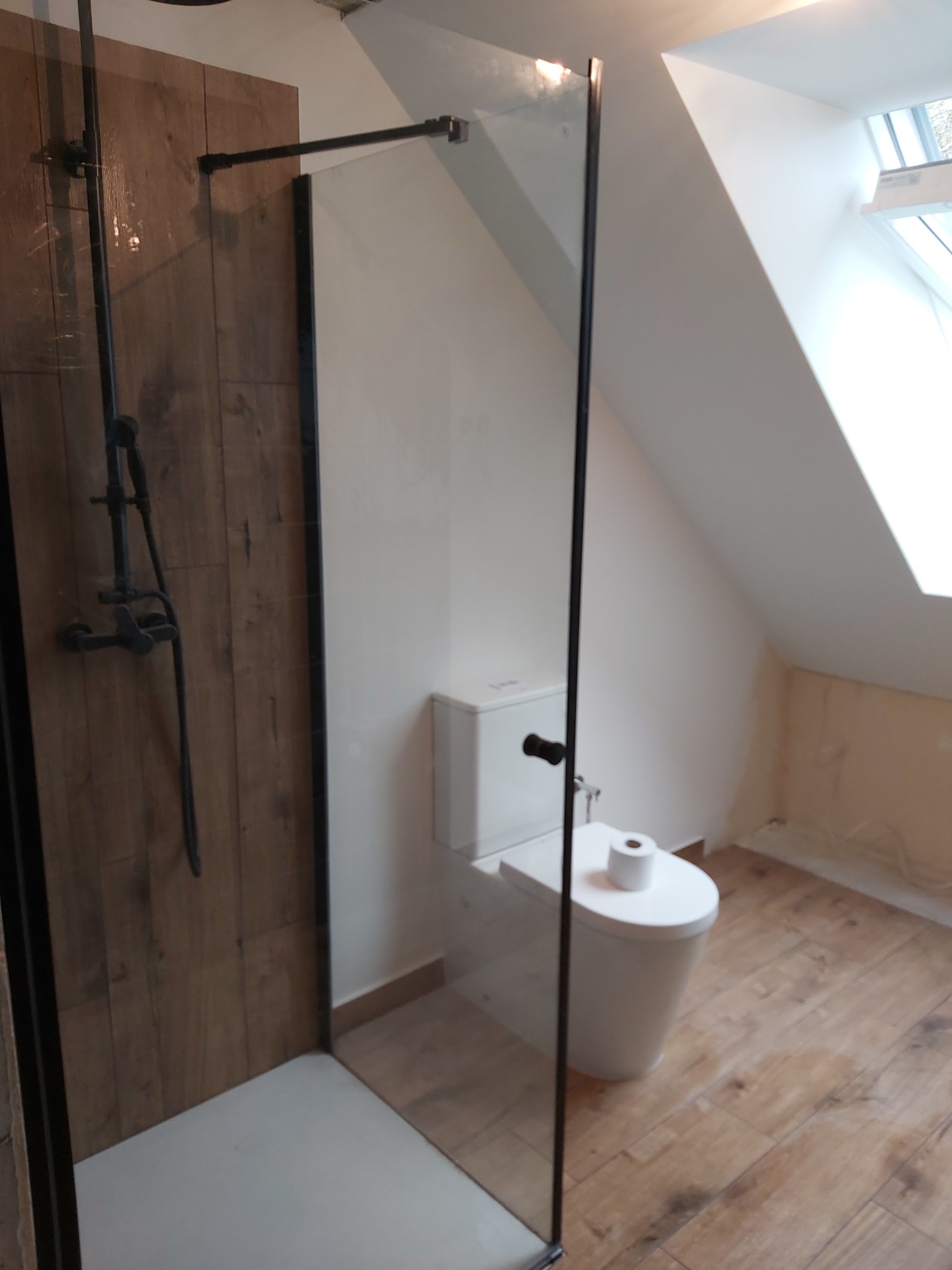 Nowoczesna łazienka na poddaszu z drewnopodobnymi płytkami, kabiną prysznicową typu walk-in z czarnymi profilami i deszczownicą w stylu retro, oraz toaletą z rolką papieru.