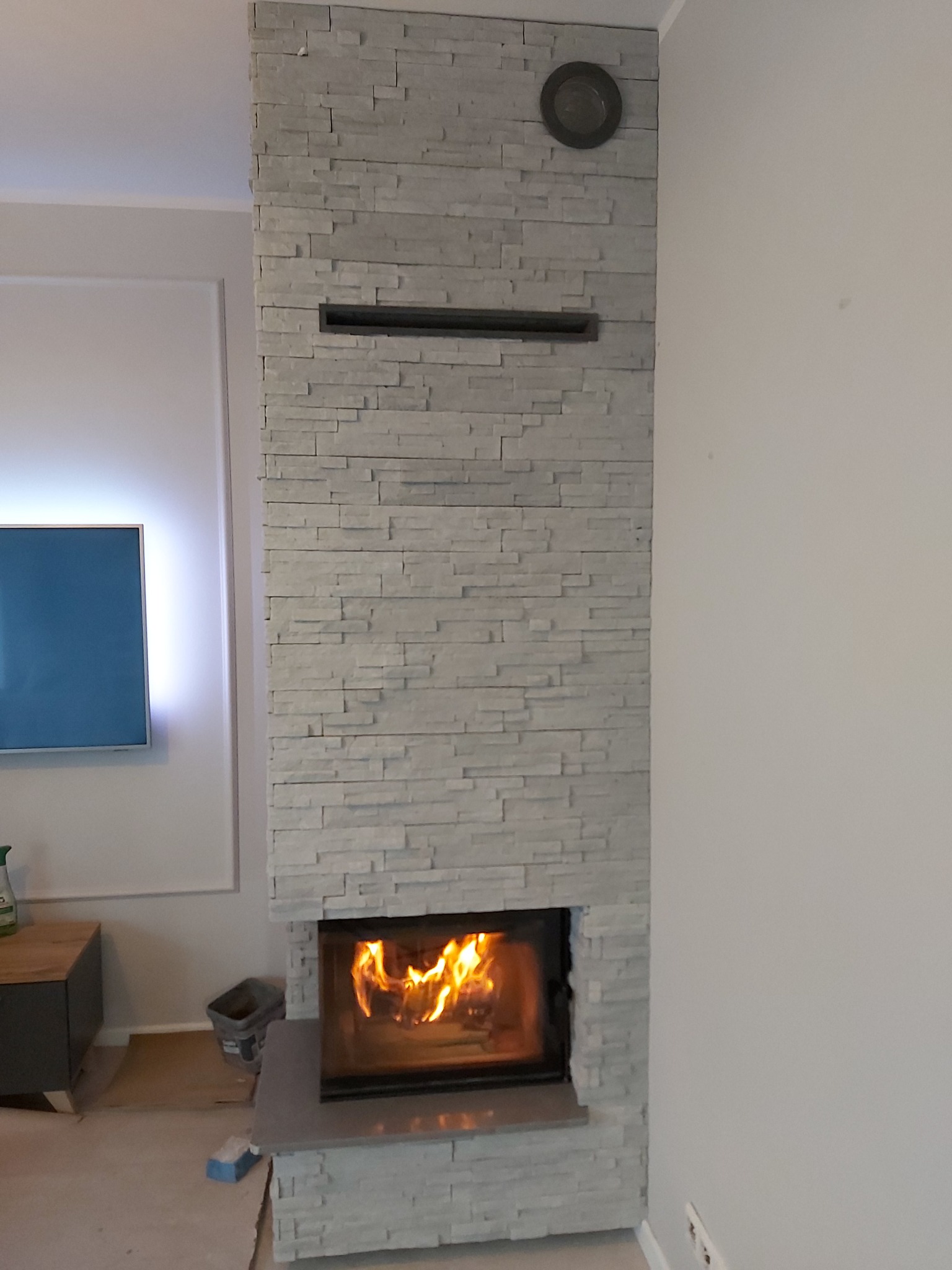 Nowoczesny kominek obłożony szarą kamienną okładziną, z widocznym paleniskiem i płomieniami, w tle ściana z telewizorem i fragment podłogi z elementami po remoncie.