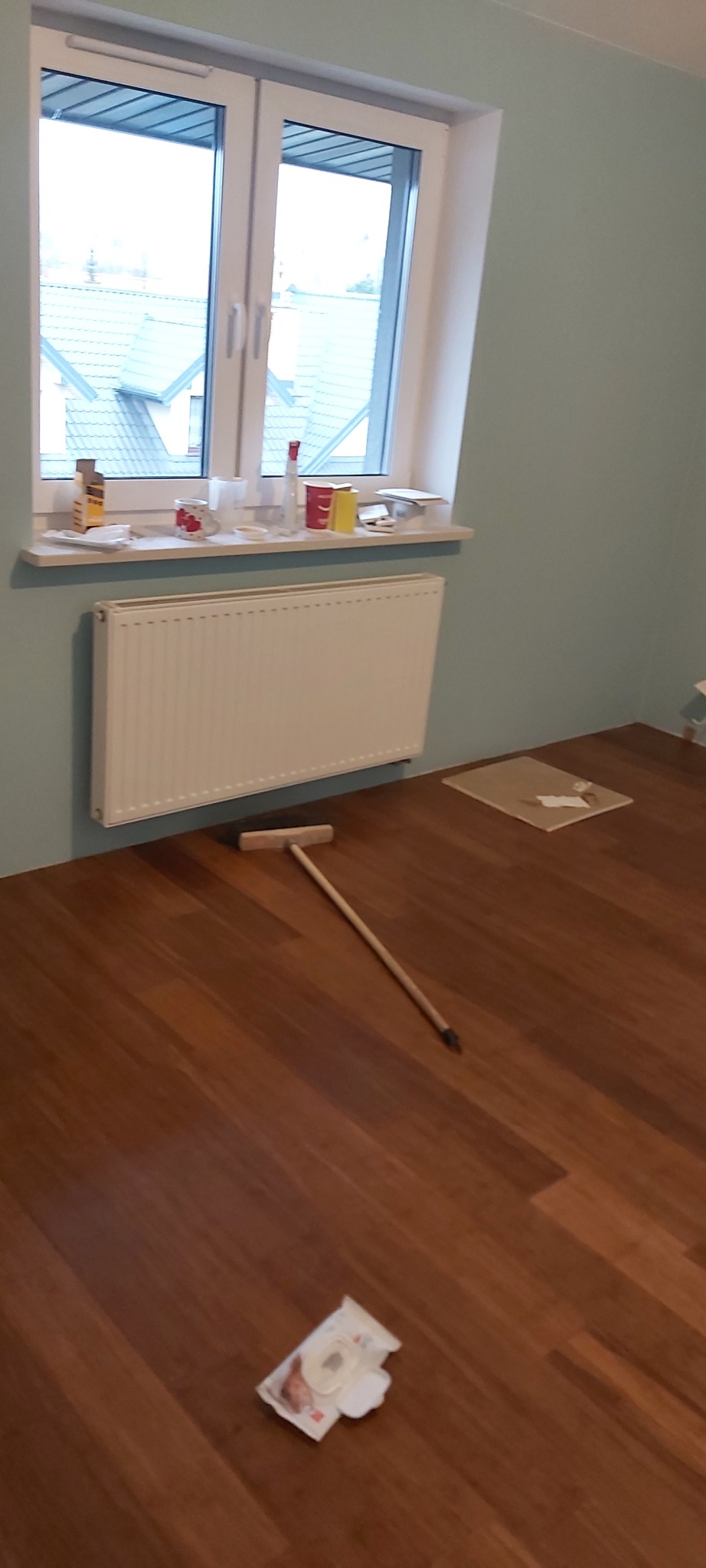 Pokój po położeniu drewnopodobnych paneli podłogowych, widoczna listwa przypodłogowa, grzejnik, okno z parapetem, oraz narzędzie do rozprowadzania kleju i resztki opakowania po kleju.