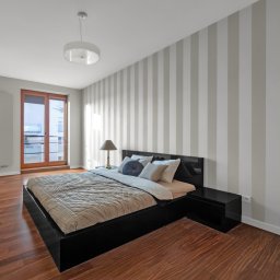 White Swan Estate - Nowoczesna sypialnia z łóżkiem na niskiej, czarnej ramie, parkietem z ciemnego drewna i ścianą w szaro-białe pasy, oświetlona lampką nocną z abażurem i lampą sufitową.