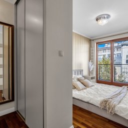 White Swan Estate - Jasna sypialnia z balkonem i drewnianą podłogą, widoczna przez otwarte drzwi, z lustrem w złotej ramie po lewej stronie i szafą wnękową.