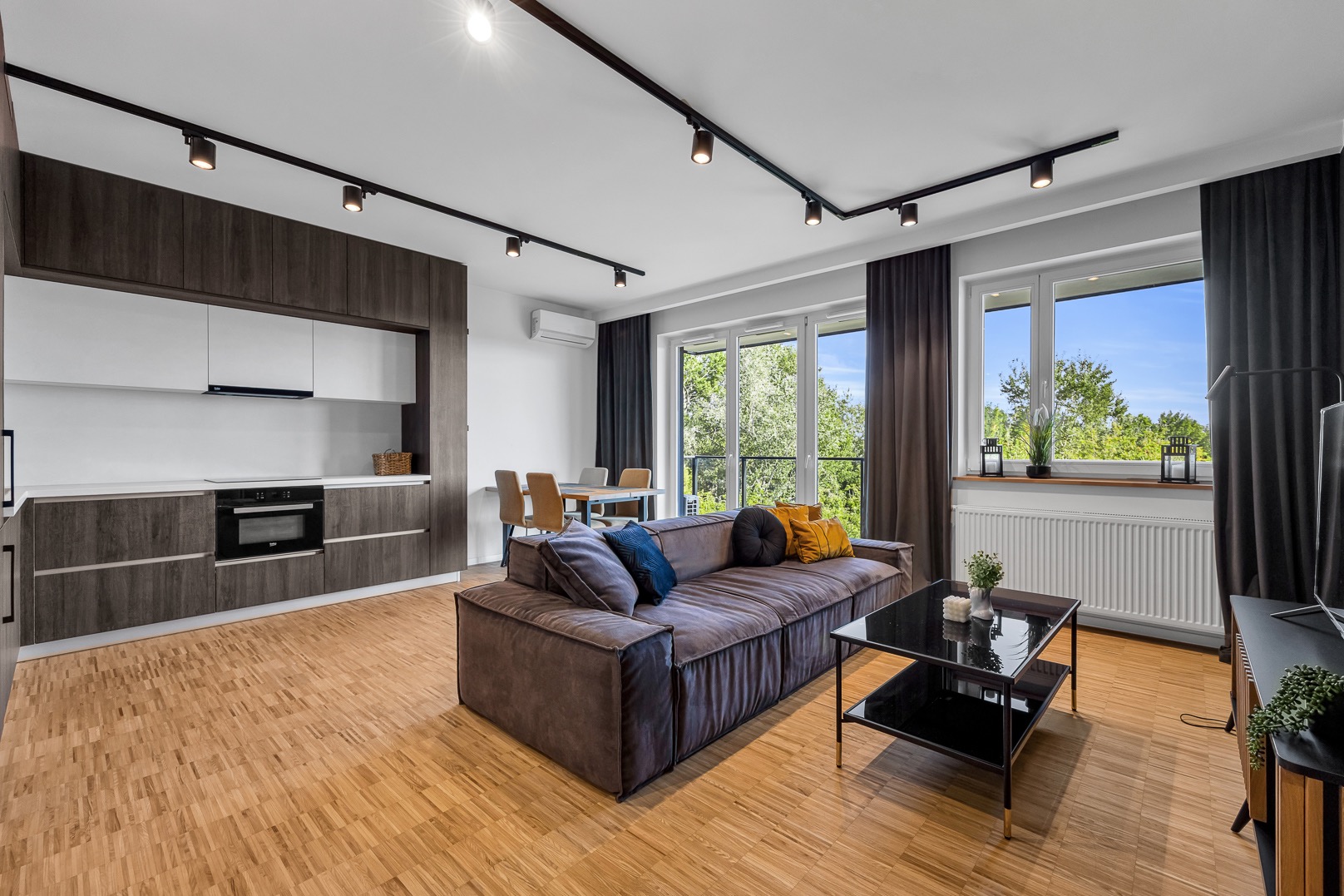 Nowoczesny salon z aneksem kuchennym, brązowa sofa z kolorowymi poduszkami, czarny stolik kawowy i parkiet w jodełkę.