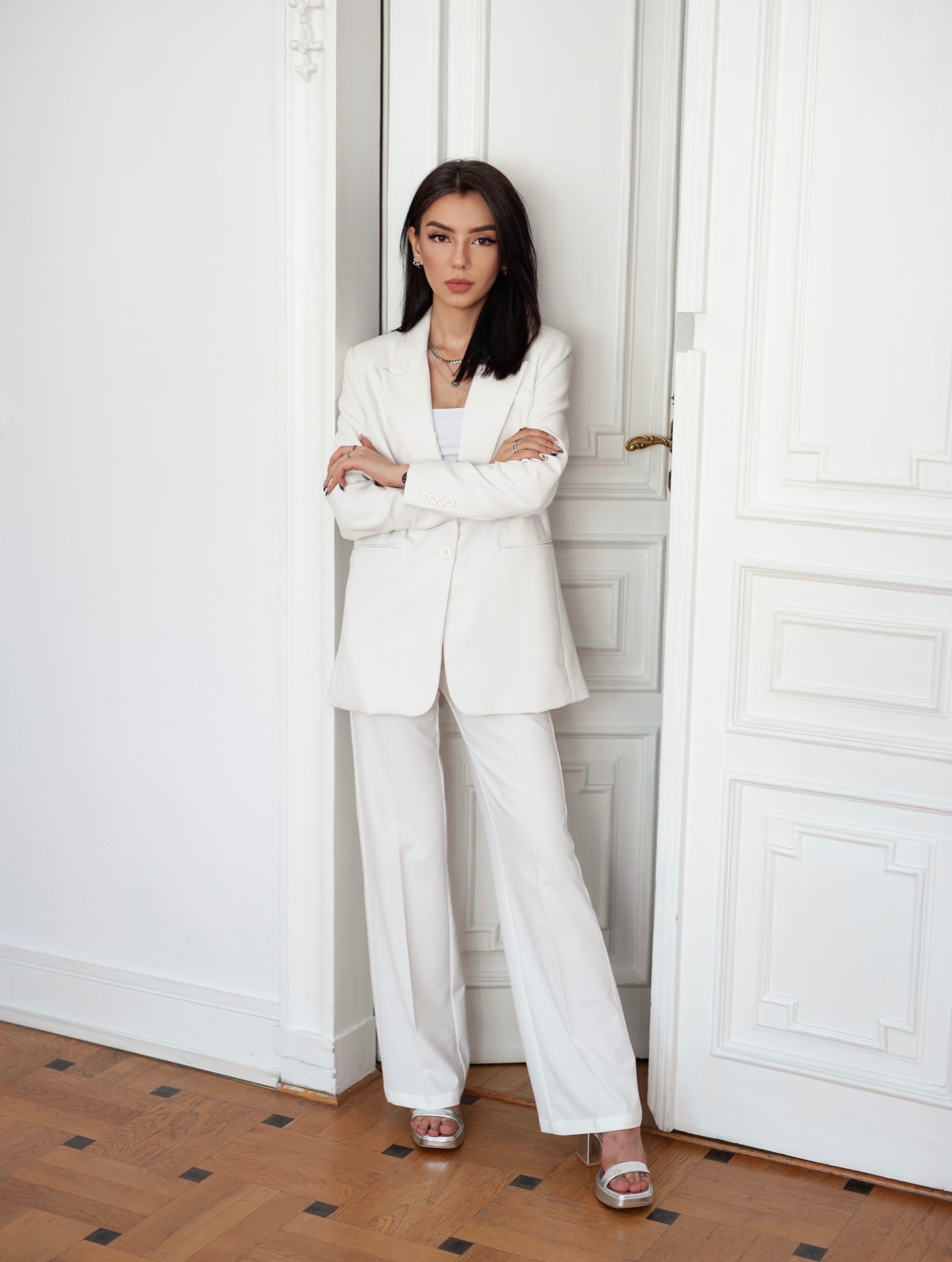 Kobieta w białym garniturze stoi oparta o białe, ozdobne drzwi w jasnym wnętrzu z parkietem w jodełkę.