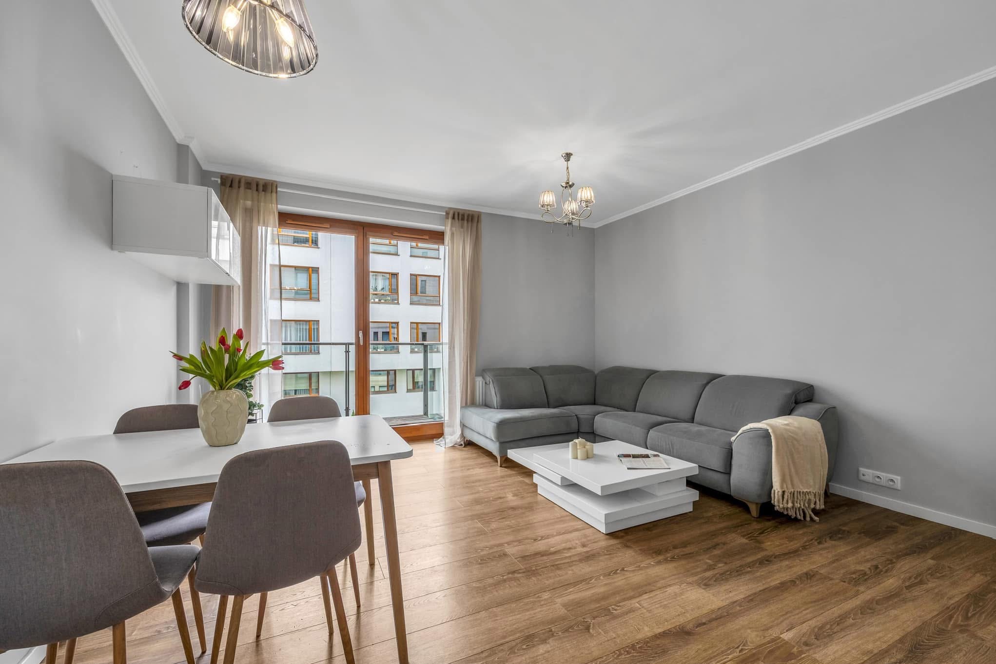 Jasne wnętrze salonu z szarą sofą narożną, białym stolikiem kawowym, stołem z tulipanami i widokiem na balkon.
