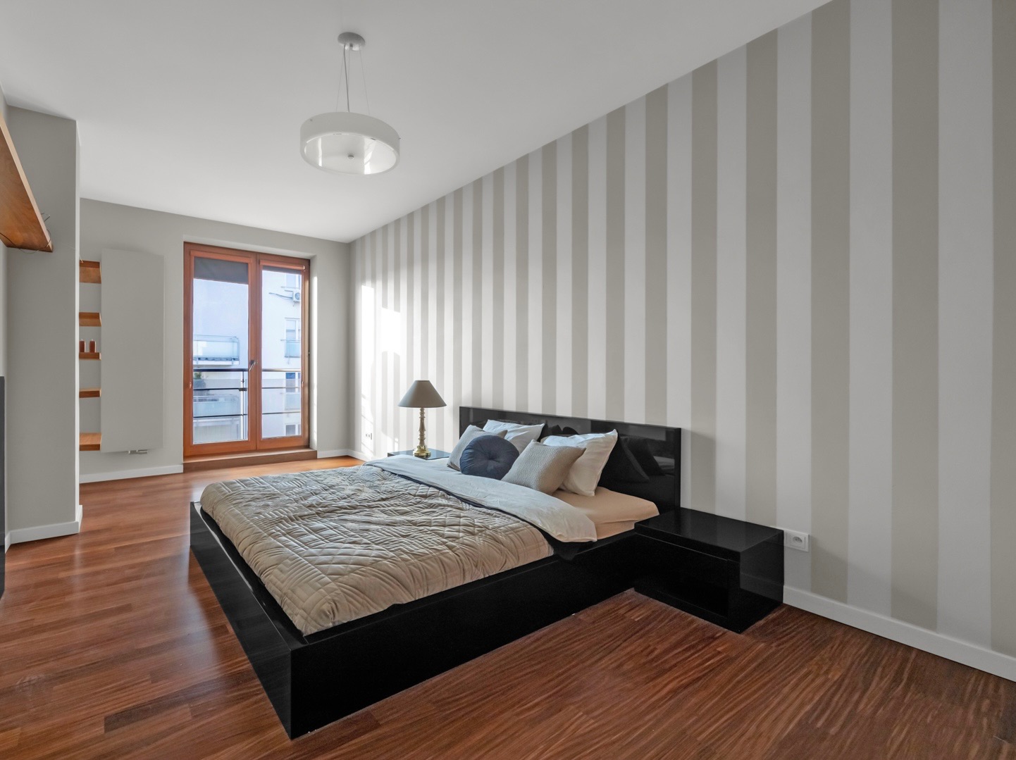 Nowoczesna sypialnia z łóżkiem na niskiej, czarnej ramie, parkietem z ciemnego drewna i ścianą w szaro-białe pasy, oświetlona lampką nocną z abażurem i lampą sufitową.