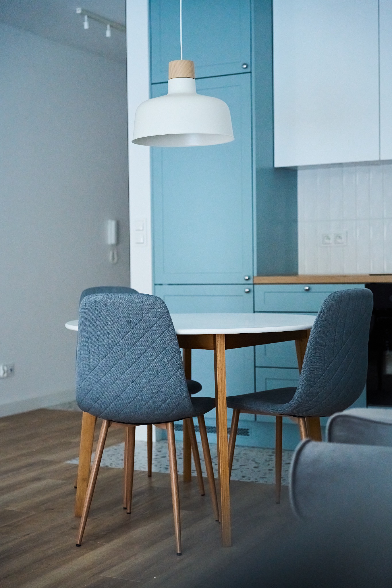 Jasne wnętrze kuchni z okrągłym białym stołem i szarymi krzesłami z drewnianymi nogami, oświetlone wiszącą lampą z drewnianym elementem, na tle błękitnych szafek kuchennych.