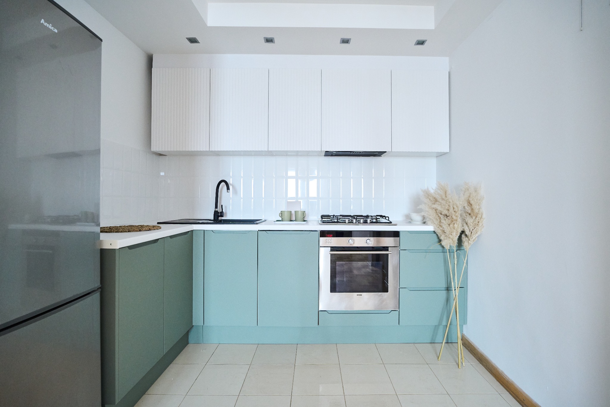 Nowoczesna kuchnia z zielonymi szafkami dolnymi i białymi górnymi, chromowanym piekarnikiem, czarną armaturą, białymi płytkami na ścianie i dekoracyjnymi trawami pampasowymi.