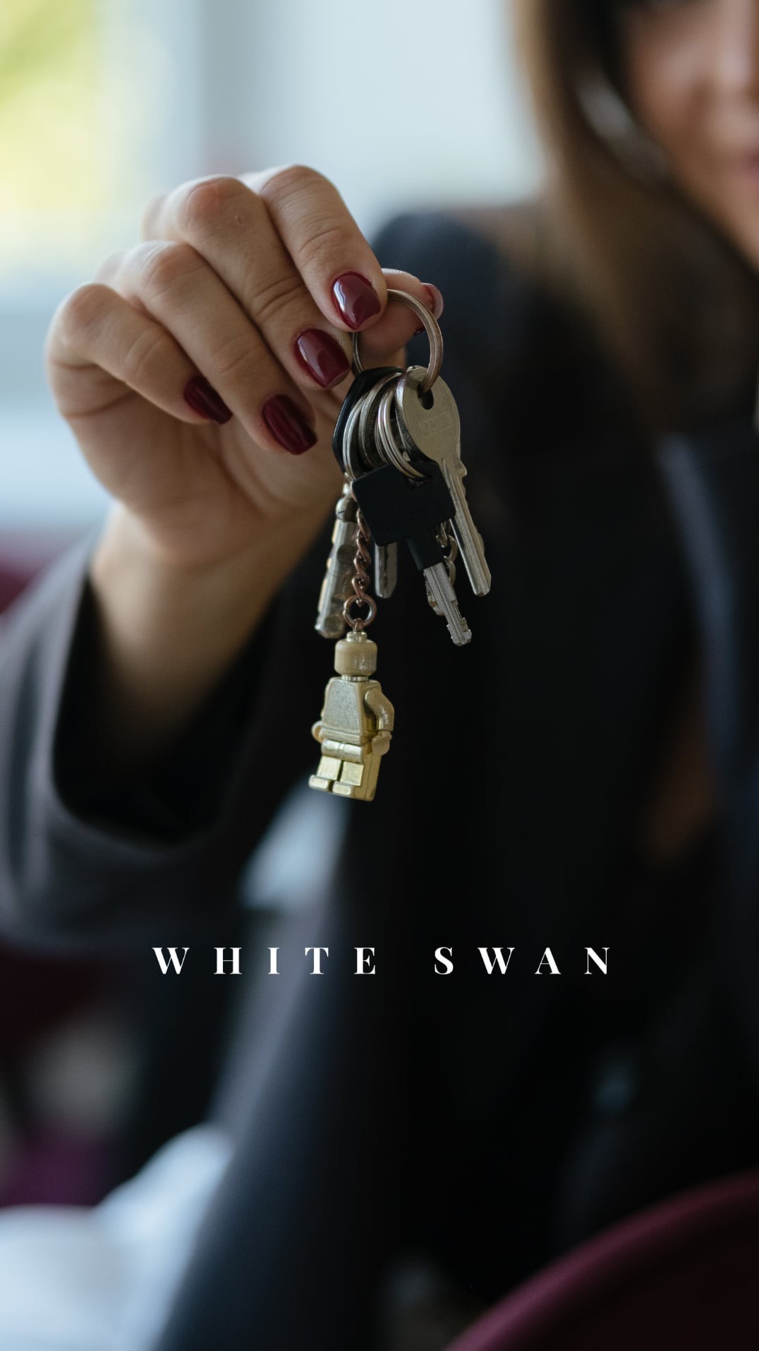 Dłoń kobiety z bordowymi paznokciami trzyma pęk kluczy z breloczkiem w kształcie złotej figurki Lego na tle okna.