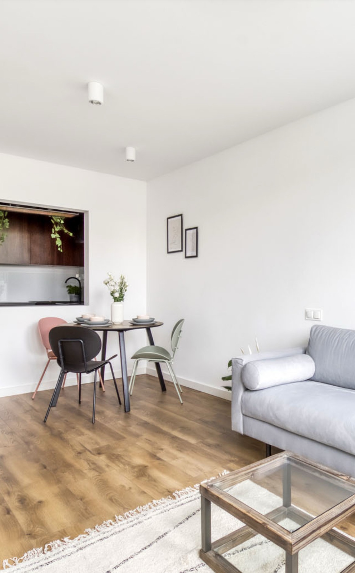 Jasne wnętrze mieszkania z drewnianą podłogą, okrągłym stołem i krzesłami w różnych kolorach, szarą sofą i szklanym stolikiem kawowym na wzorzystym dywanie.