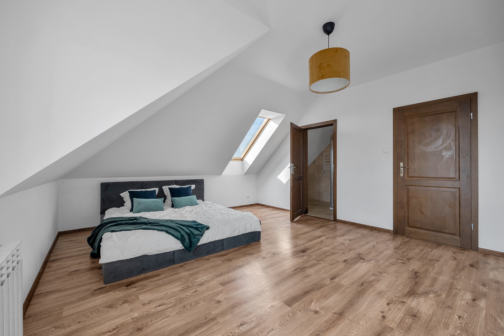 Jasna sypialnia na poddaszu z drewnianą podłogą, skośnym oknem dachowym, szarą ramą łóżka z niebieskimi poduszkami i zielonym pledem, oraz otwartymi drzwiami do łazienki z widocznym grzejnikiem...