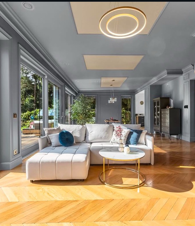 Jasny salon z parkietem w jodełkę, narożną sofą, okrągłym stolikiem kawowym i nowoczesnym oświetleniem sufitowym