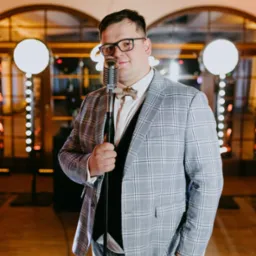 Mężczyzna w okularach i kraciastej marynarce trzymający mikrofon retro na tle dekoracji świetlnych.