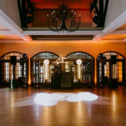 Sala balowa z parkietem, oświetleniem scenicznym i neonowym napisem 'Love & Dance' nad stanowiskiem DJ-a, widoczne głośniki i reflektory na statywach, oświetlenie w ciepłych barwach.