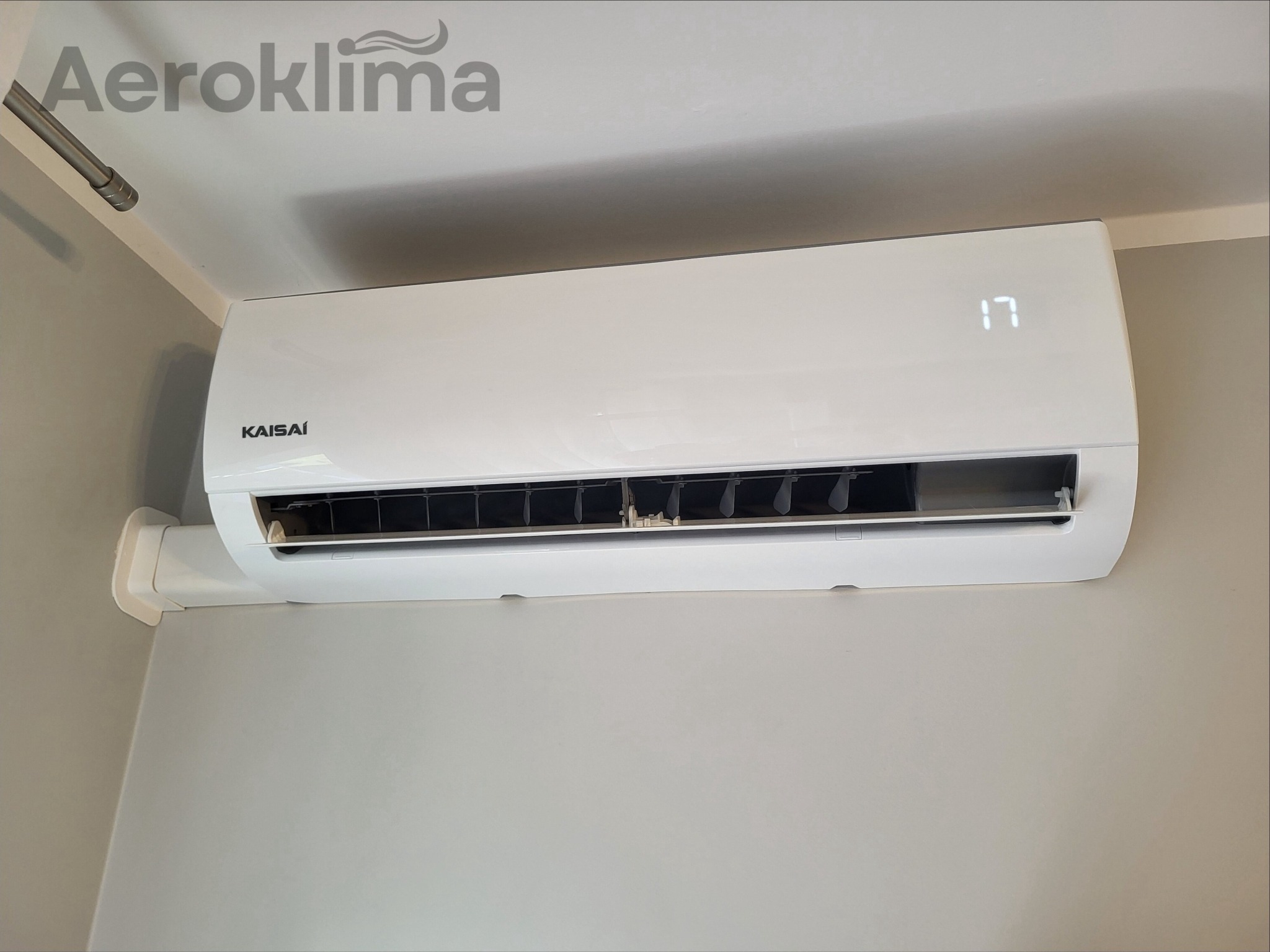 Biały klimatyzator Kaisai z wyświetlaczem temperatury 17 stopni Celsjusza, zamontowany na szarej ścianie w pomieszczeniu.