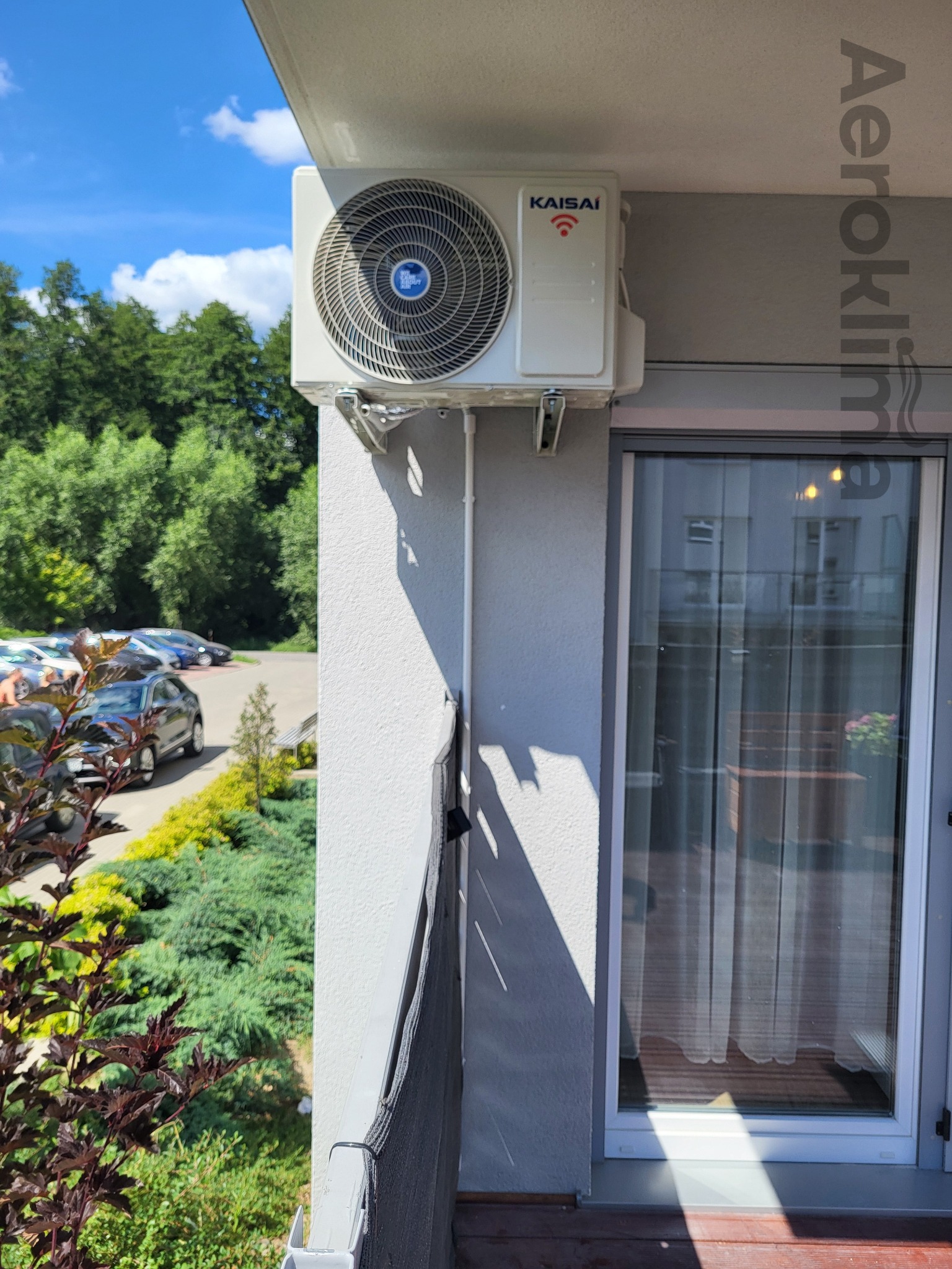 Zewnętrzna jednostka klimatyzacji Kaisai zamontowana na szarej ścianie balkonu z widokiem na parking i zieleń.