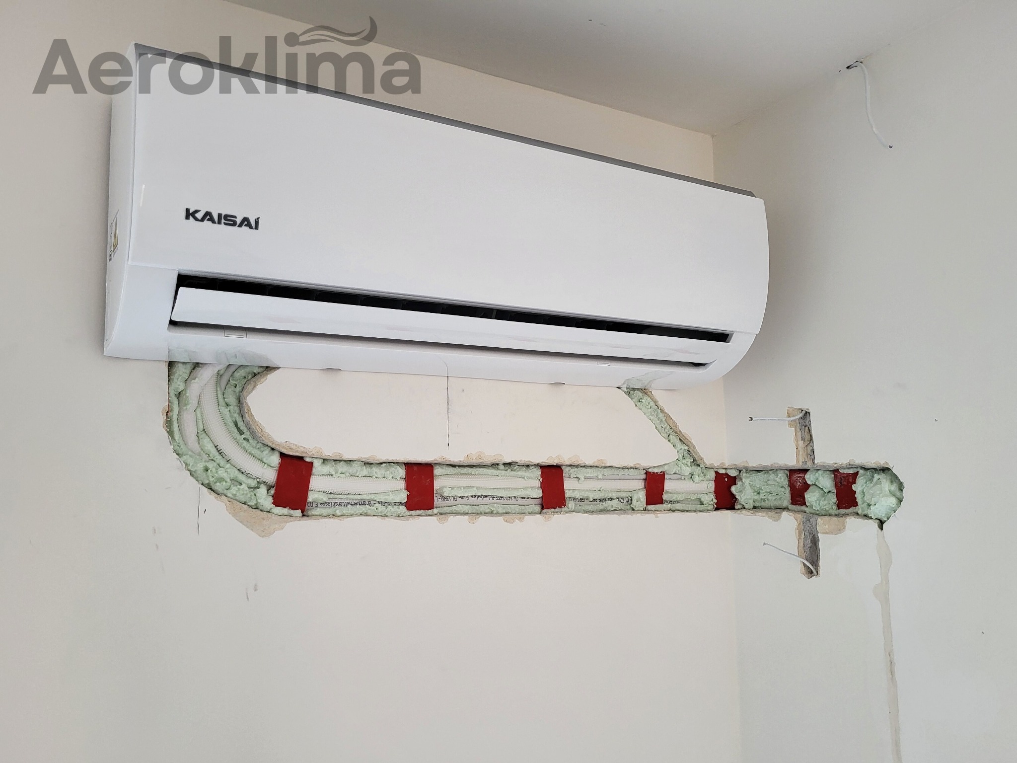 Biały klimatyzator Kaisai zamontowany na ścianie, z widocznymi przewodami chłodniczymi zabezpieczonymi pianką montażową i czerwonymi uchwytami, częściowo ukrytymi w ścianie.