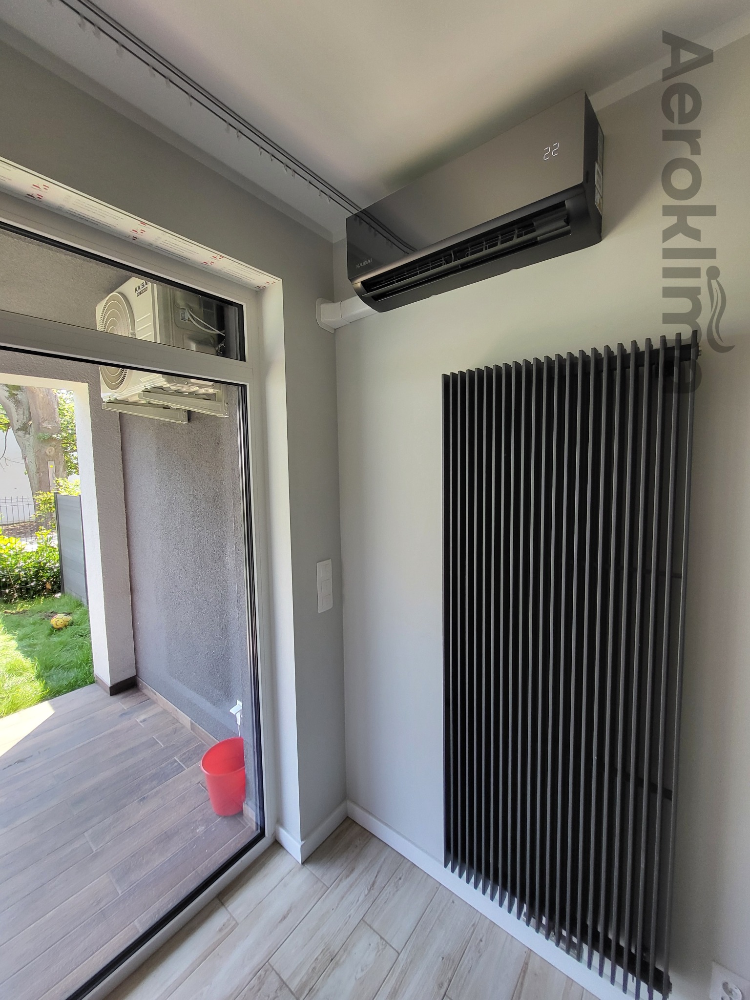 Nowoczesny, grafitowy klimatyzator ścienny z wyświetlaczem temperatury w pomieszczeniu z ciemnym grzejnikiem panelowym, widok przez otwarte drzwi balkonowe na zewnątrz.