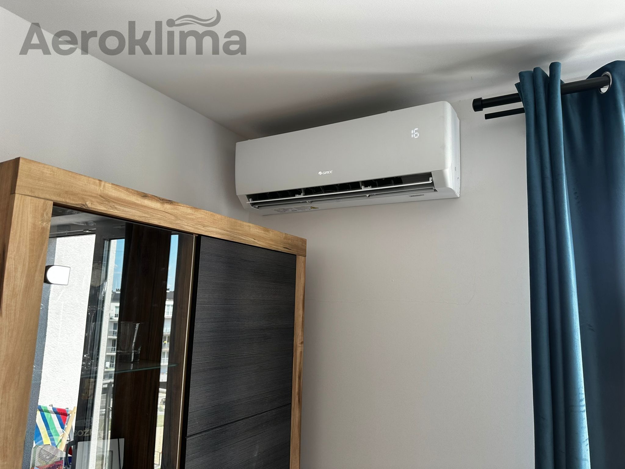Biały klimatyzator marki Gree z wyświetloną temperaturą 16 stopni, zamontowany na ścianie w pokoju, obok ciemnego regału i zasłony w kolorze morskim.