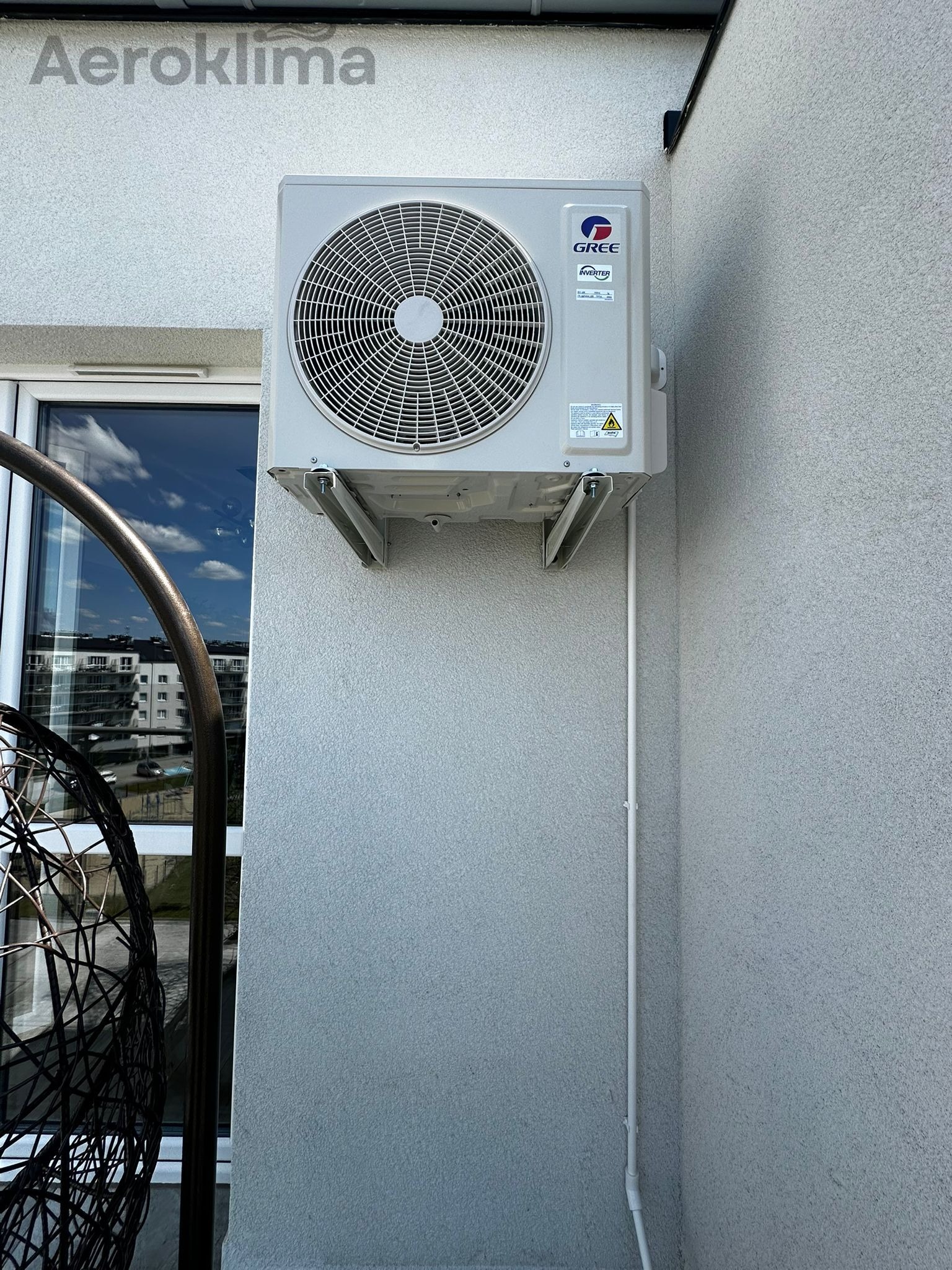 Zewnętrzna jednostka klimatyzacji Gree zamontowana na ścianie budynku obok okna, widoczny fragment wiszącego fotela.