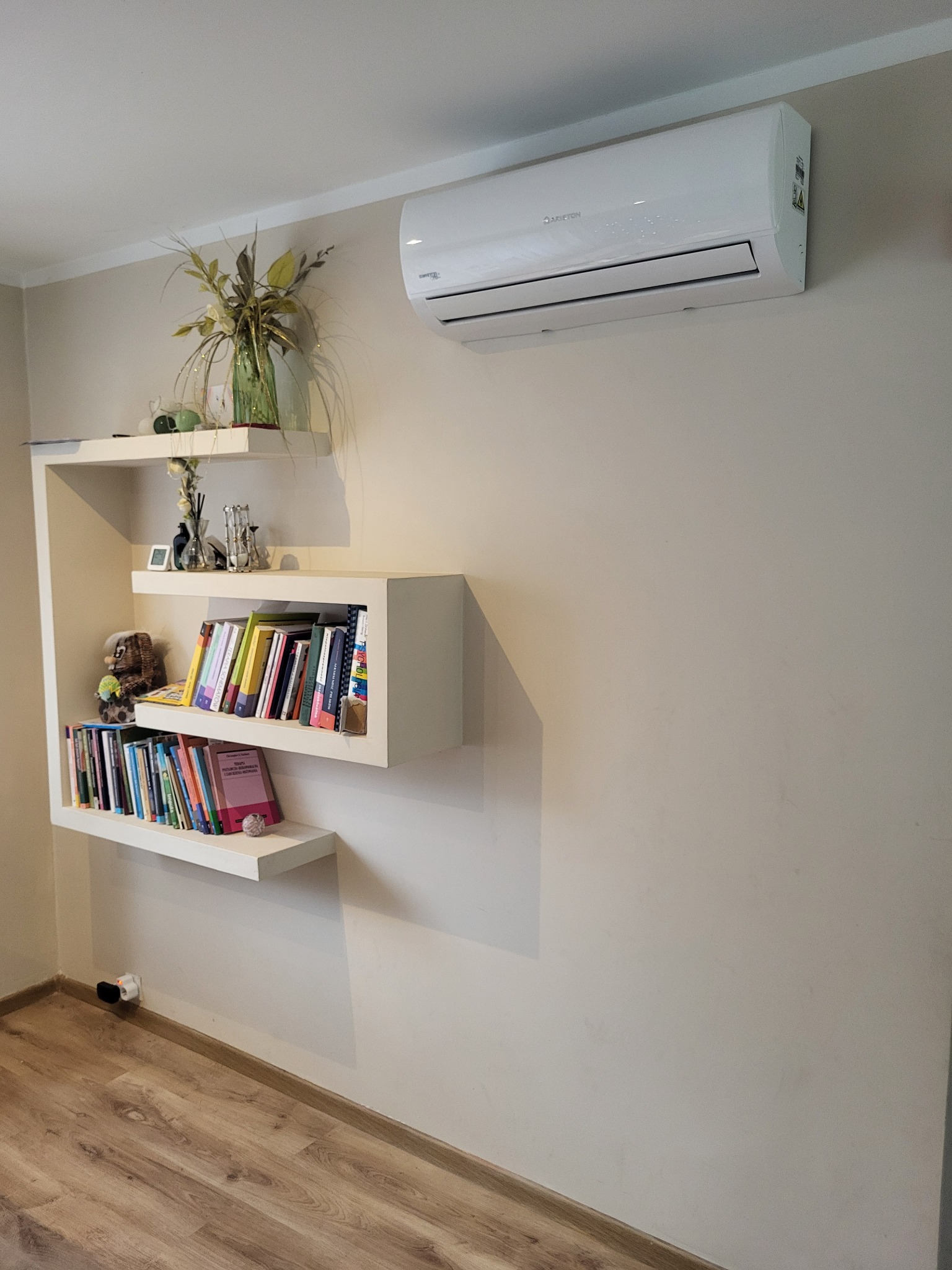Biały klimatyzator Ariston zamontowany na ścianie nad nowoczesnym regałem z książkami i dekoracjami w jasnym wnętrzu.