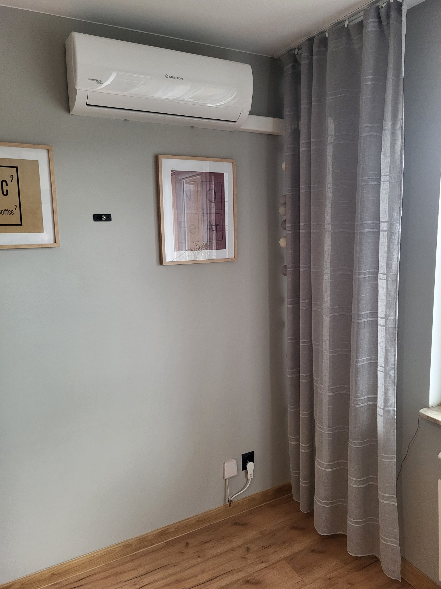 Biała jednostka klimatyzacyjna Ariston zamontowana na szarej ścianie w pokoju z dwoma obrazami i zasłonami w tle.