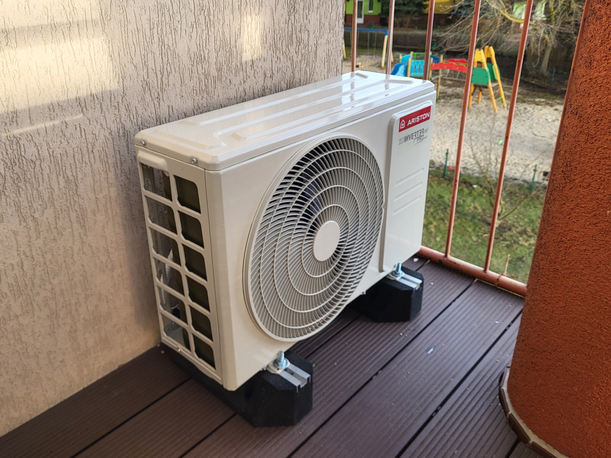 Zewnętrzna jednostka klimatyzacji Ariston Inverter Pro zamontowana na balkonie z drewnianą podłogą i widokiem na plac zabaw.
