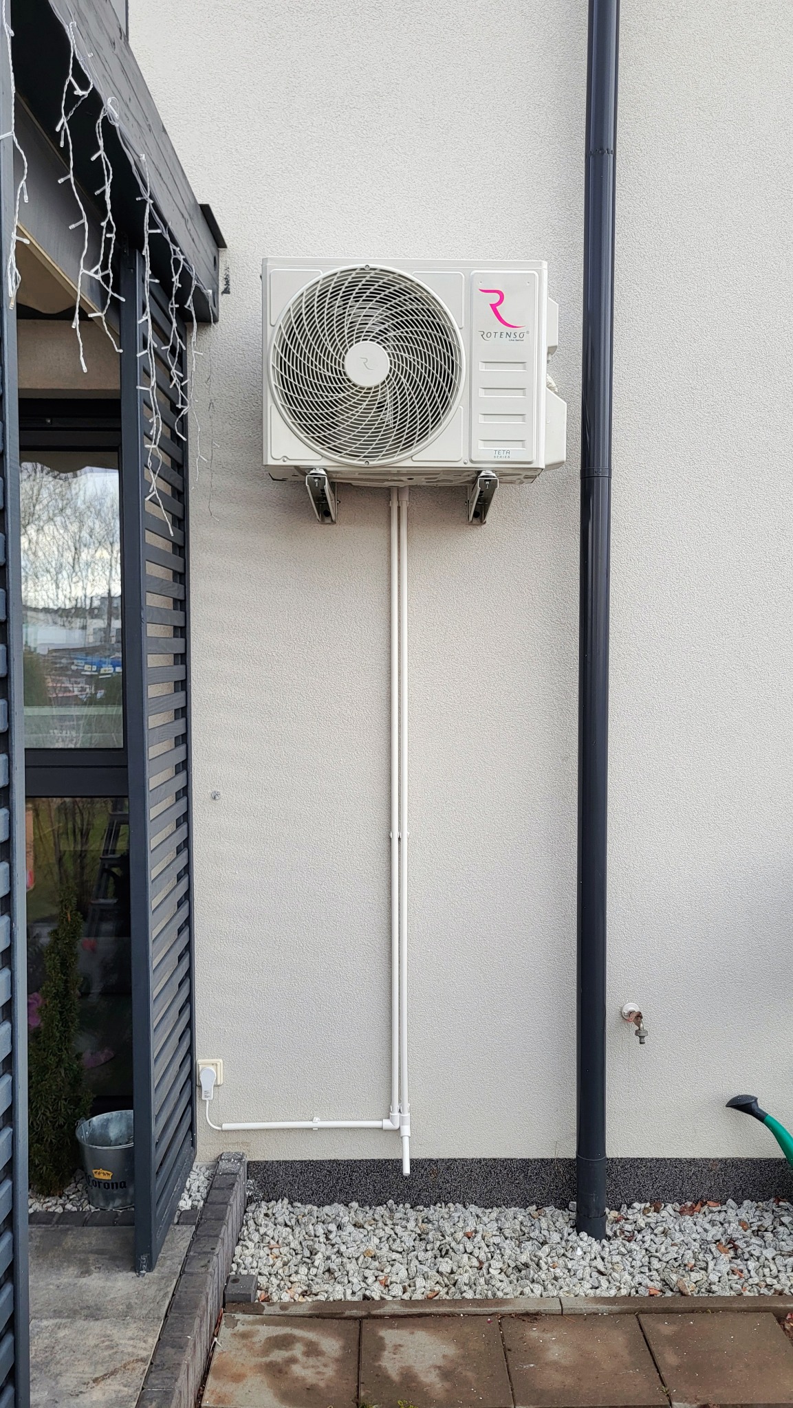 Zewnętrzna jednostka klimatyzacji Rotenso zamontowana na ścianie domu, widoczne rury instalacyjne, elewacja o fakturze baranka i fragment systemu odprowadzania wody deszczowej.