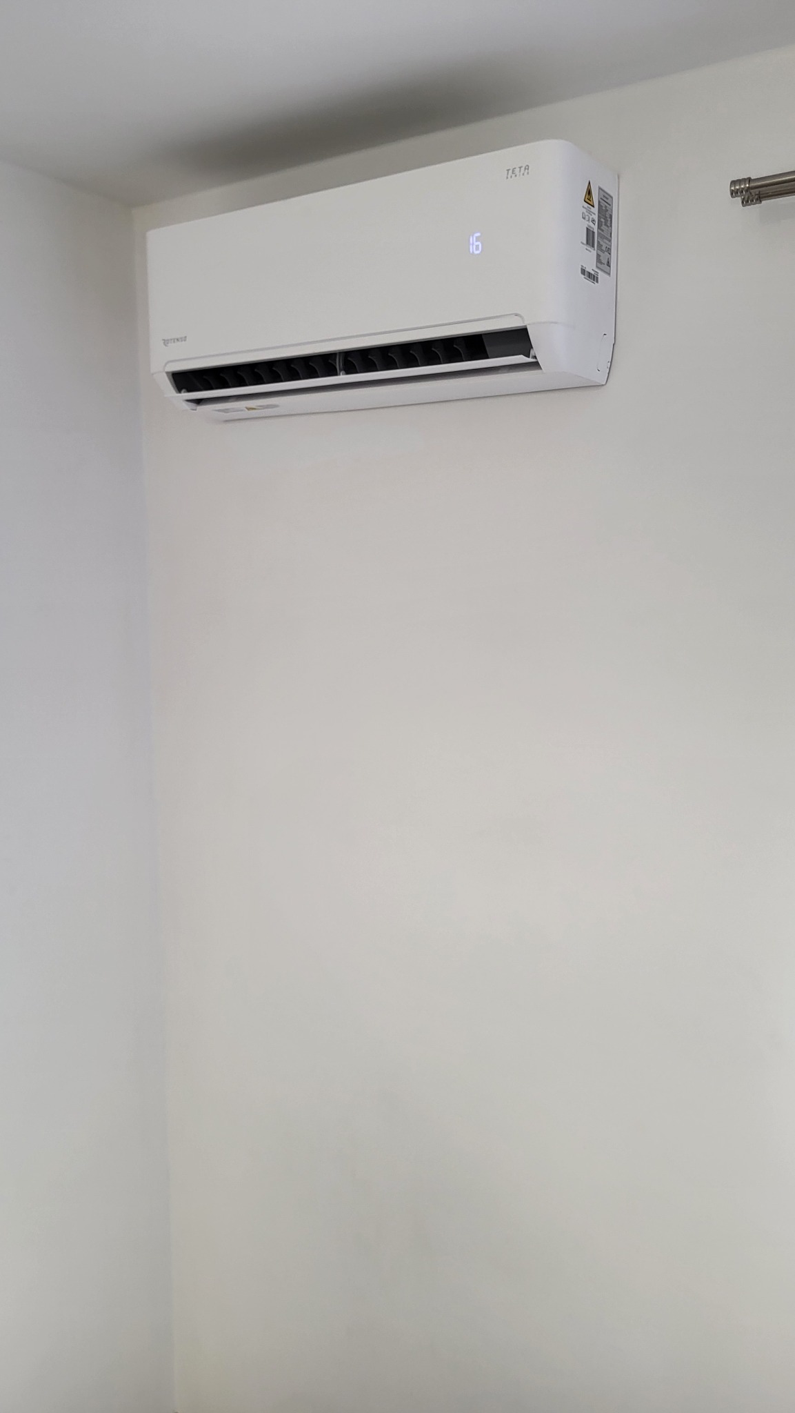 Biała klimatyzacja TATA zamontowana na białej ścianie, wyświetlacz pokazuje temperaturę 16 stopni, widoczna rura na górze po prawej stronie.