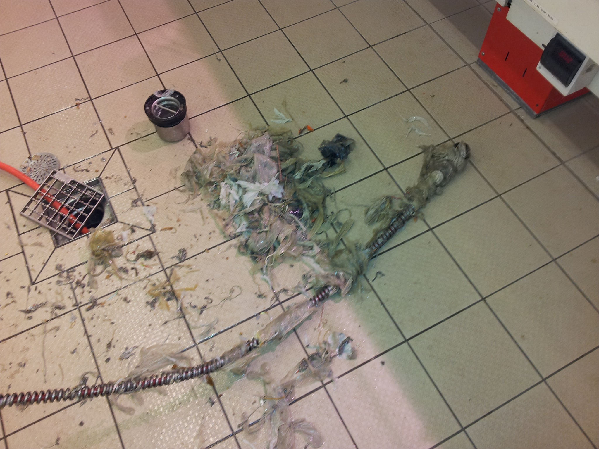 Udrożniona kratka ściekowa na kafelkach, obok leżą narzędzia i wyciągnięte zanieczyszczenia, w tle widoczny fragment urządzenia.