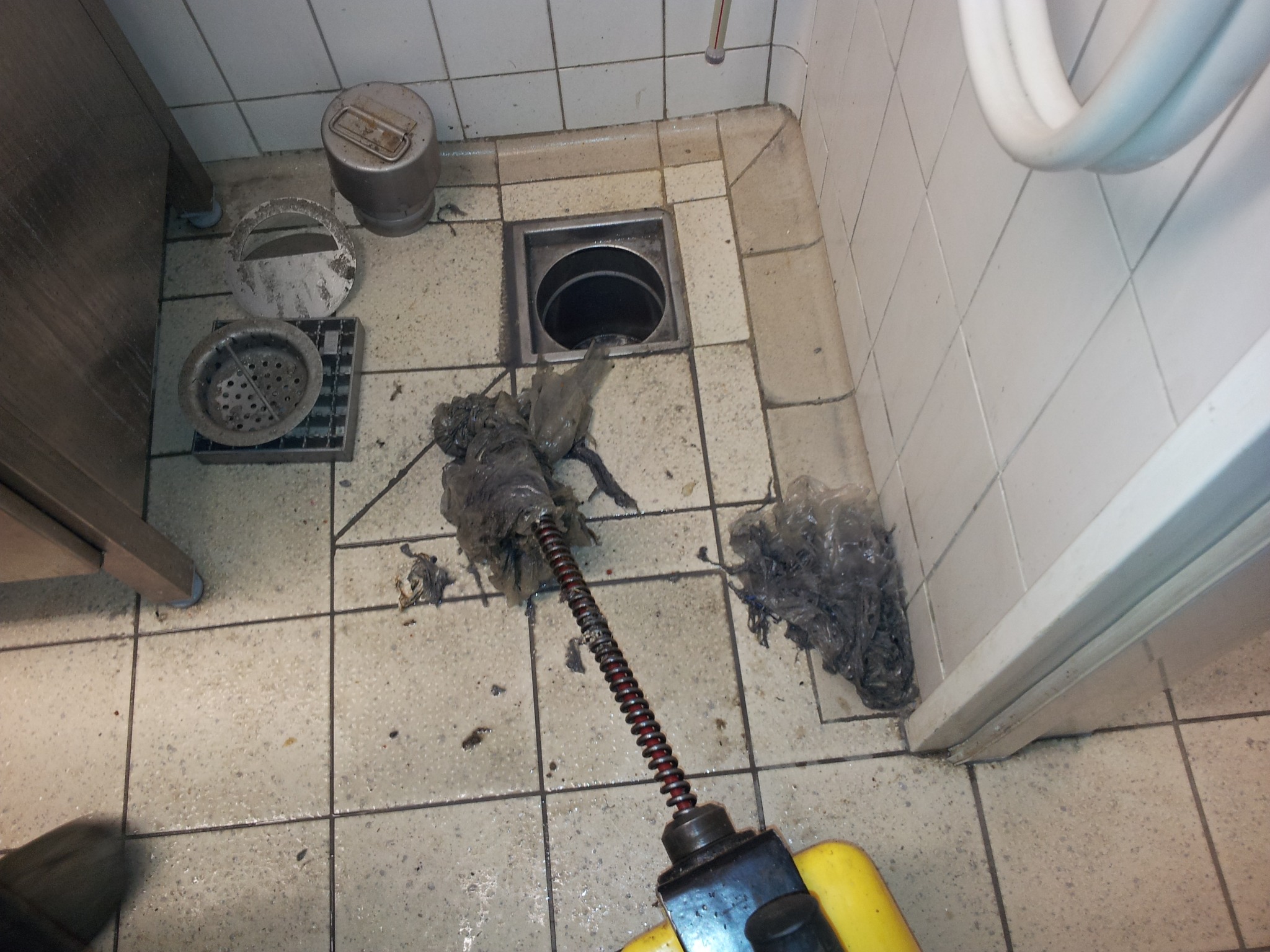 Udrażnianie odpływu podłogowego w łazience z użyciem elektrycznej sprężyny, widoczne wyciągnięte zanieczyszczenia i elementy kratki odpływowej.