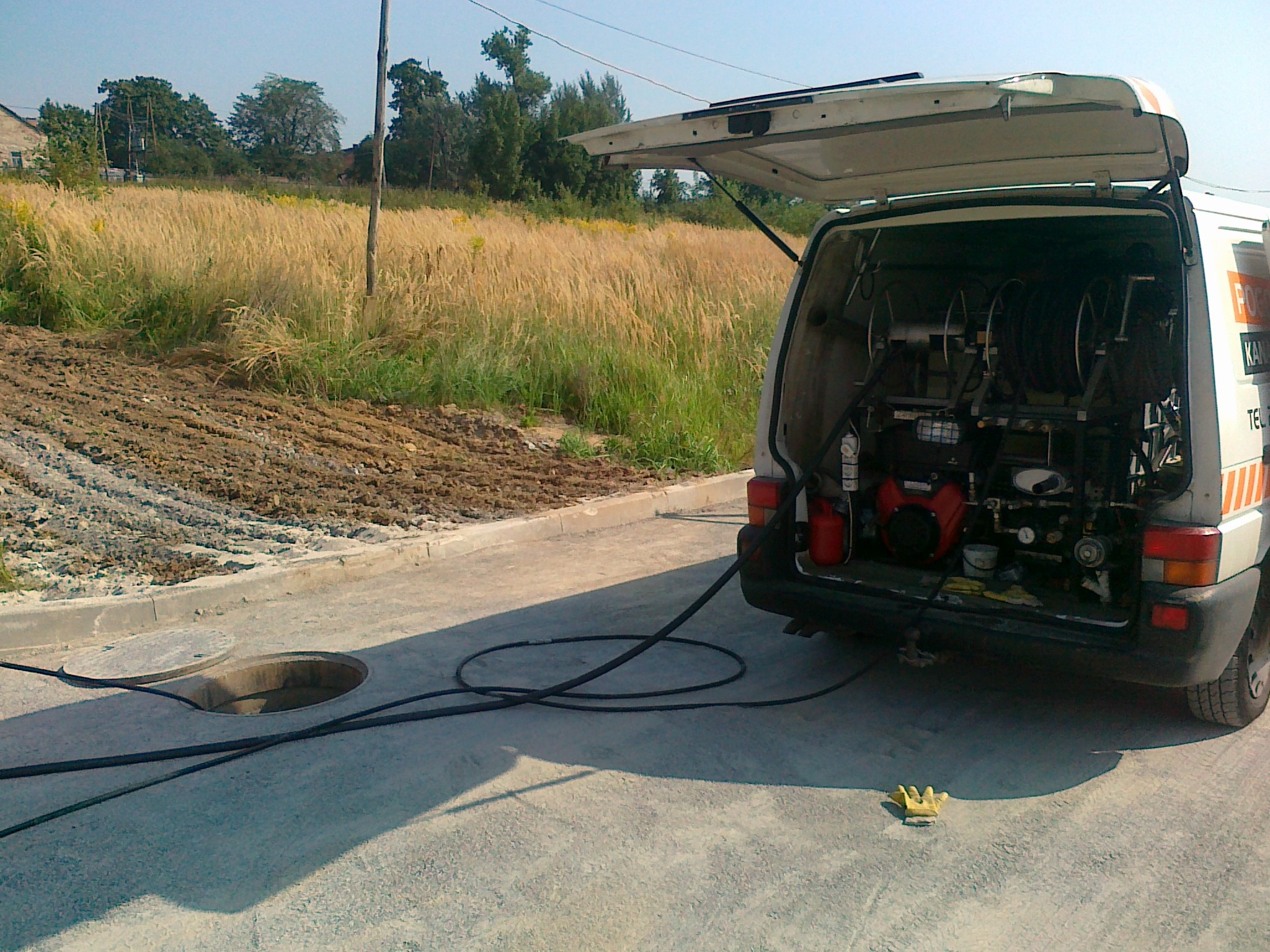 Otwarty bagażnik samochodu serwisowego z urządzeniem do udrażniania rur, z widocznym włazem kanalizacyjnym i wężem na ziemi oraz żółtą rękawicą roboczą.