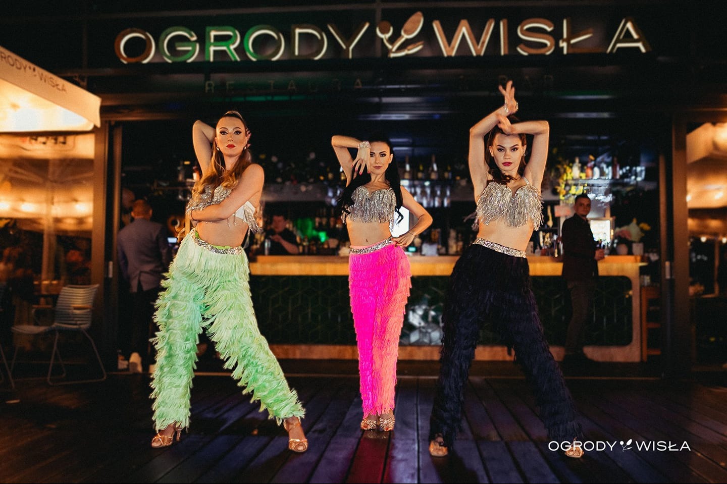 Trzy tancerki w kostiumach z frędzlami (zielone, różowe, czarne) prezentują układ taneczny na tle baru o nazwie 'Ogrody Wisła'.