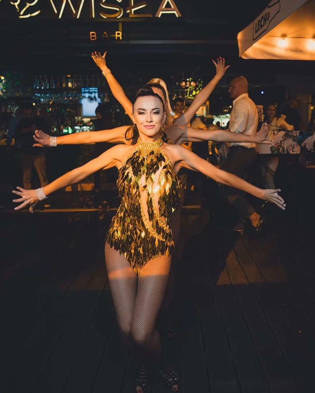 Tancerka w złotym kostiumie z elementami przypominającymi liście, na tle baru z napisem 'WISEA BAR', z rozłożonymi ramionami w towarzystwie innych tancerek.