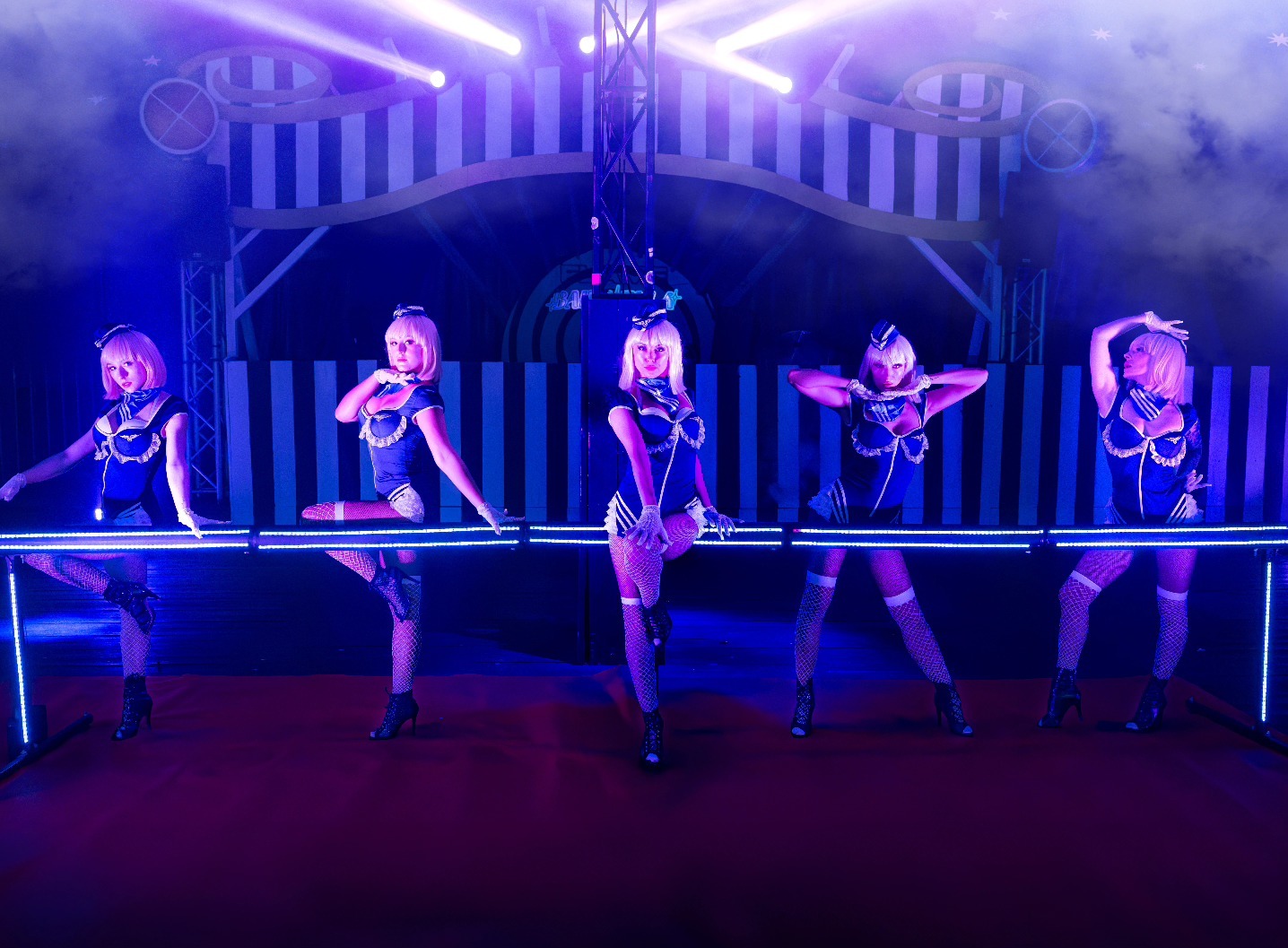 Zespół tancerek w niebieskich kostiumach i siatkowych pończochach wykonuje układ taneczny na scenie z neonowym oświetleniem i dekoracjami w tle.