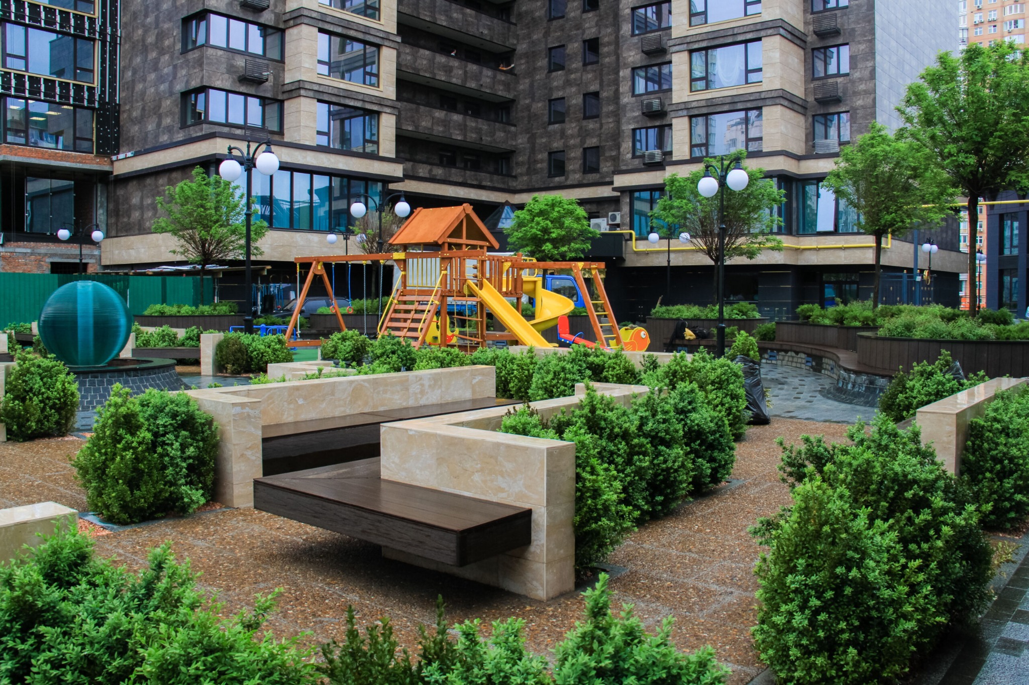 Nowoczesny plac zabaw z drewnianą konstrukcją i zjeżdżalnią, otoczony bujną zielenią krzewów i niskich drzew, na tle wielopiętrowego budynku mieszkalnego z ciemną elewacją, oświetlony kulistymi...