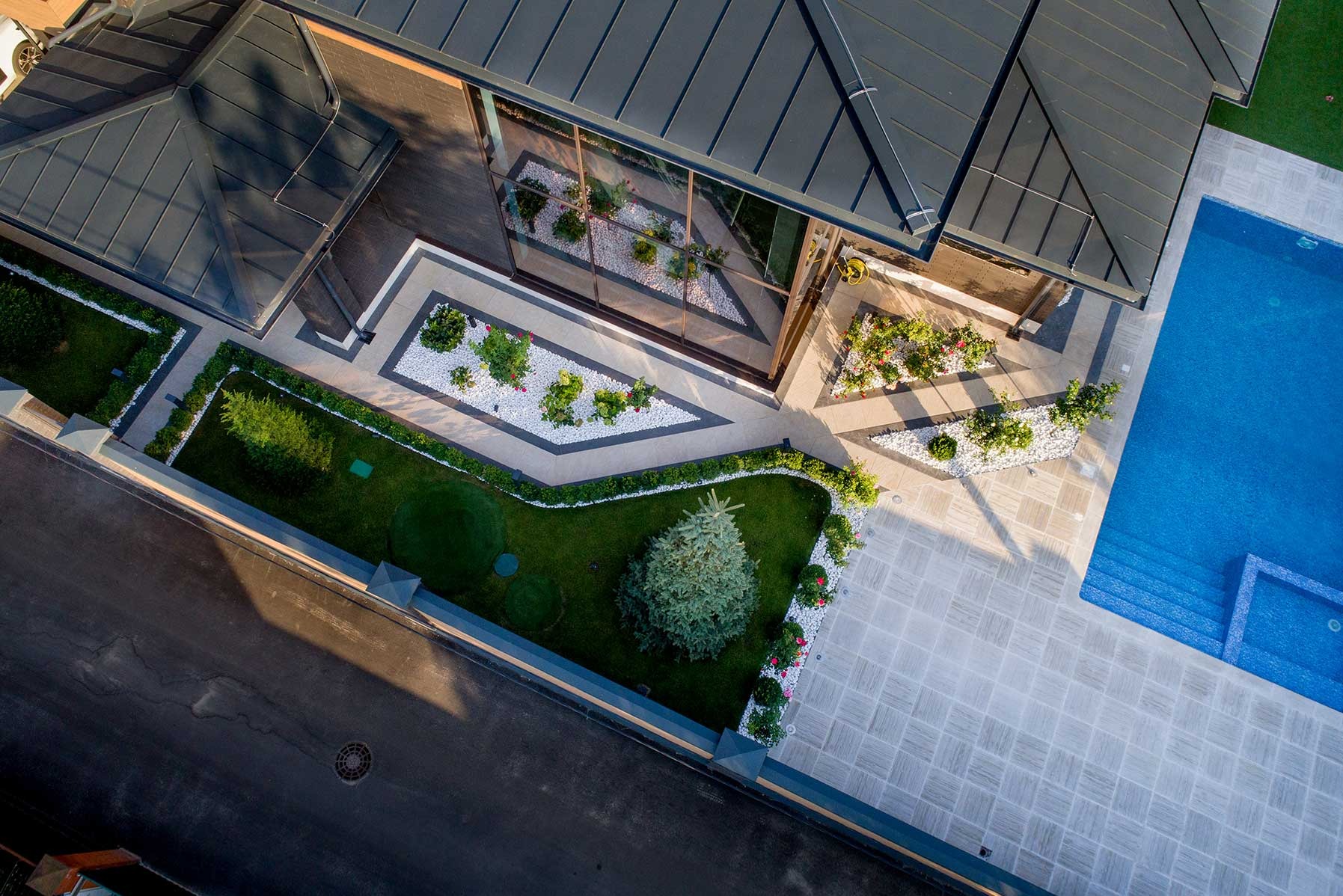 Widok z góry na nowoczesny ogród z basenem, trawnikiem, białymi kamieniami dekoracyjnymi i geometrycznymi rabatami, otoczony kostką brukową w odcieniach szarości i beżu, z elementami architektury...