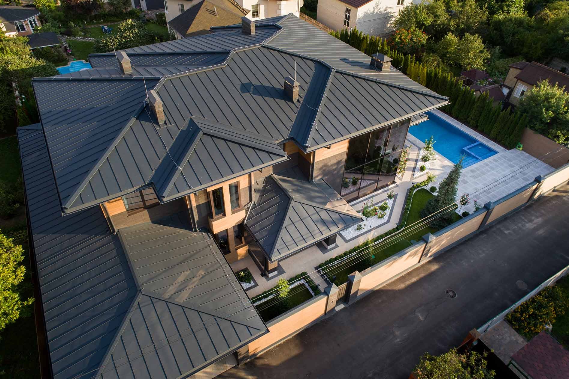 Widok z góry na luksusowy dom z szarym dachem blaszanym na rąbek stojący, dużymi oknami, basenem z jasną kostką brukową, zadbanym ogrodem z białymi kamykami i wysokim ogrodzeniem.
