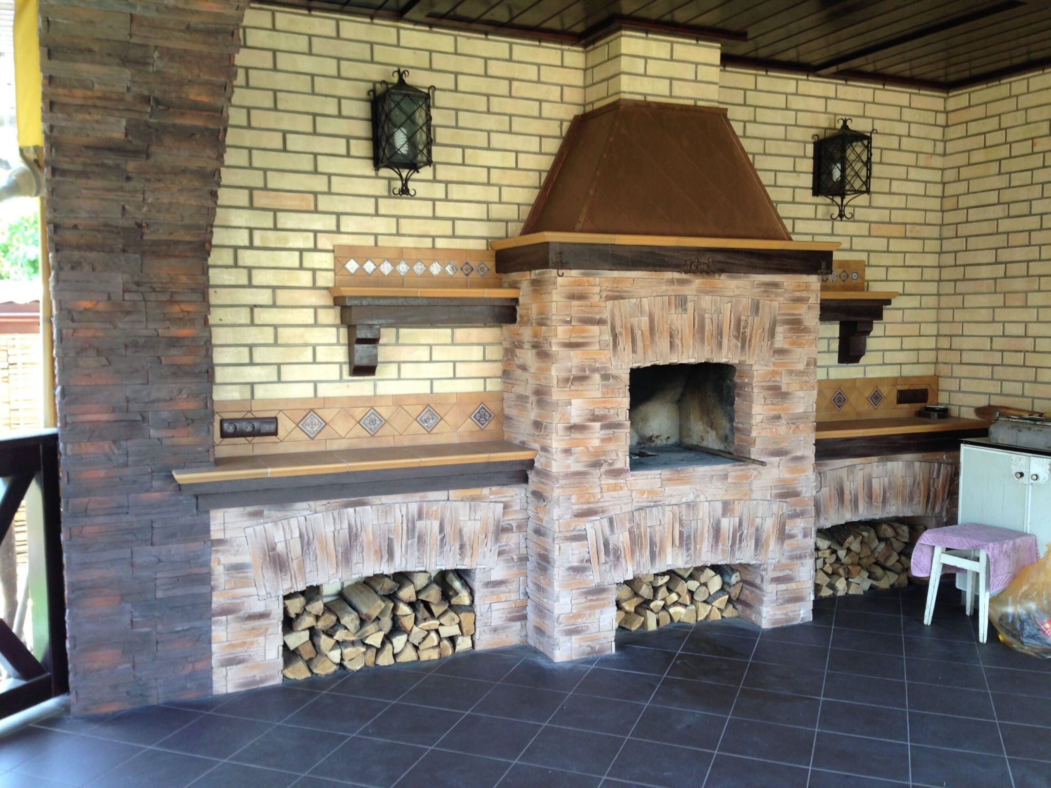 Zewnętrzny kominek z cegły i kamienia, z zapasem drewna pod spodem, oświetlony dekoracyjnymi lampami ściennymi.