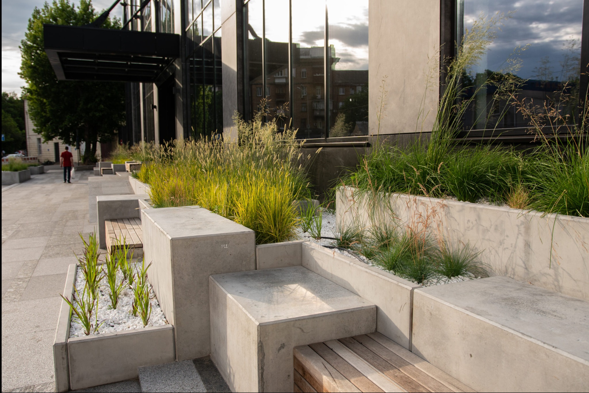 Nowoczesna przestrzeń miejska z betonowymi donicami, w których rosną trawy ozdobne i młode rośliny, wysypane białym grysem, połączone z siedziskami z drewnianych desek, na tle budynku...