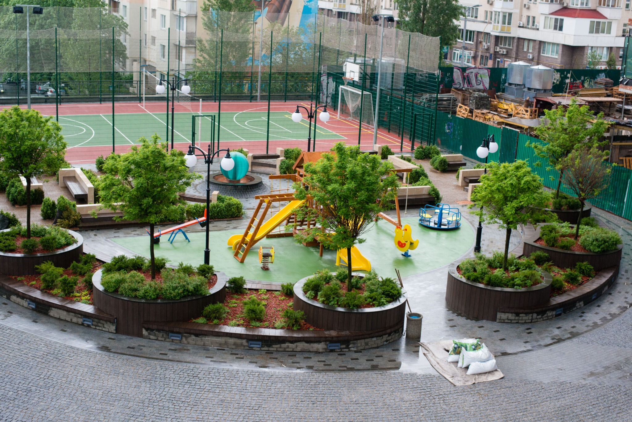 Widok z góry na plac zabaw z elementami zieleni w dużych donicach, w tle boisko sportowe i budynki mieszkalne.