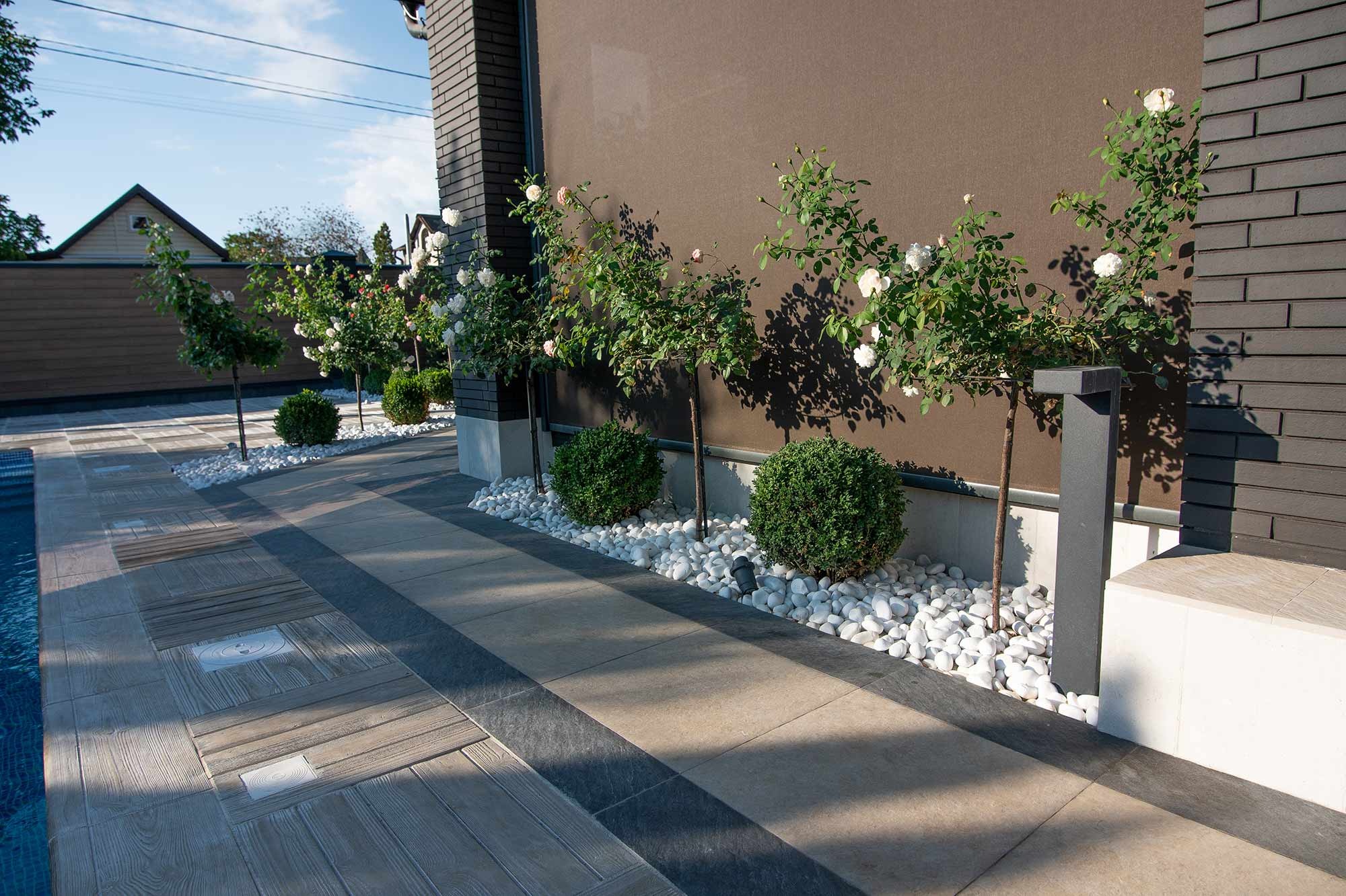 Ogród z białymi różami na pniu, obok ścieżki z kostki brukowej i białych kamieni, z systemem oświetlenia ogrodowego