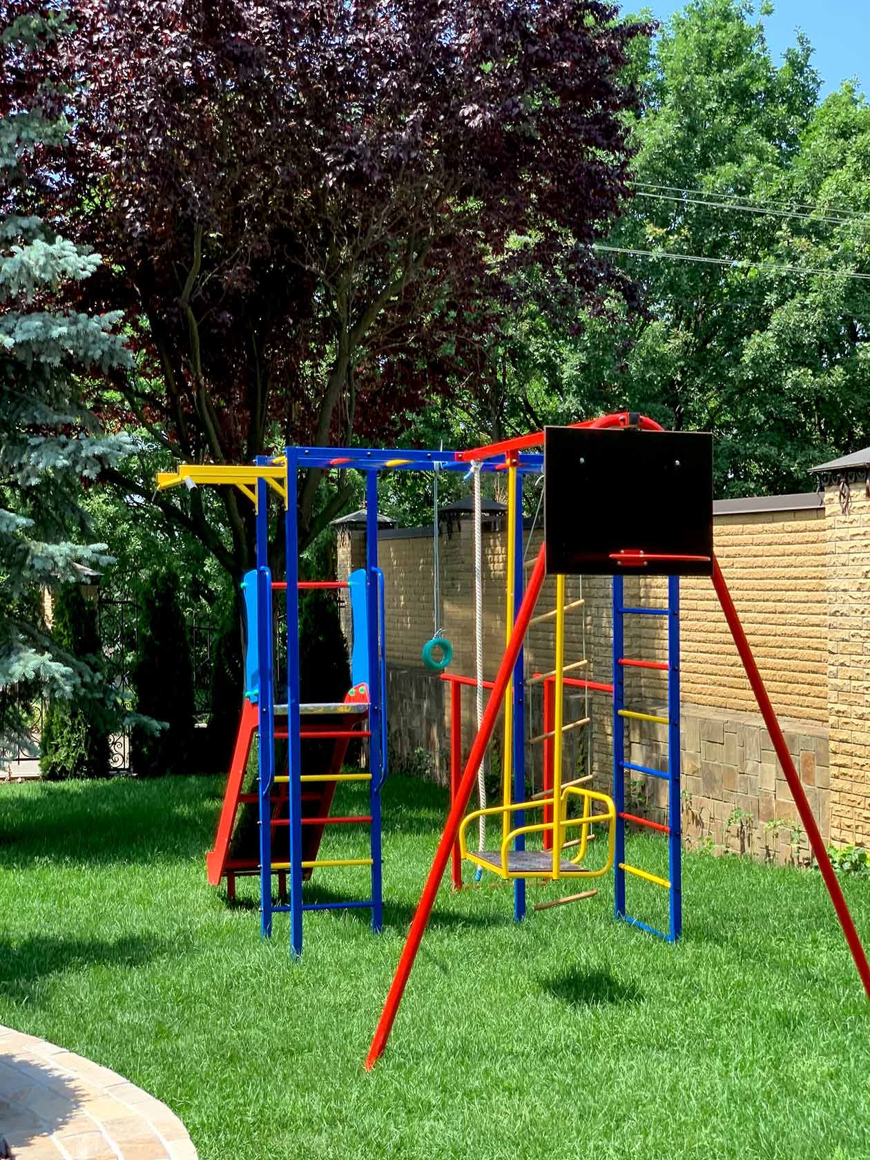 Kolorowy plac zabaw z huśtawką, zjeżdżalnią i koszem do koszykówki na zadbanym trawniku, w tle drzewa i mur z piaskowca.