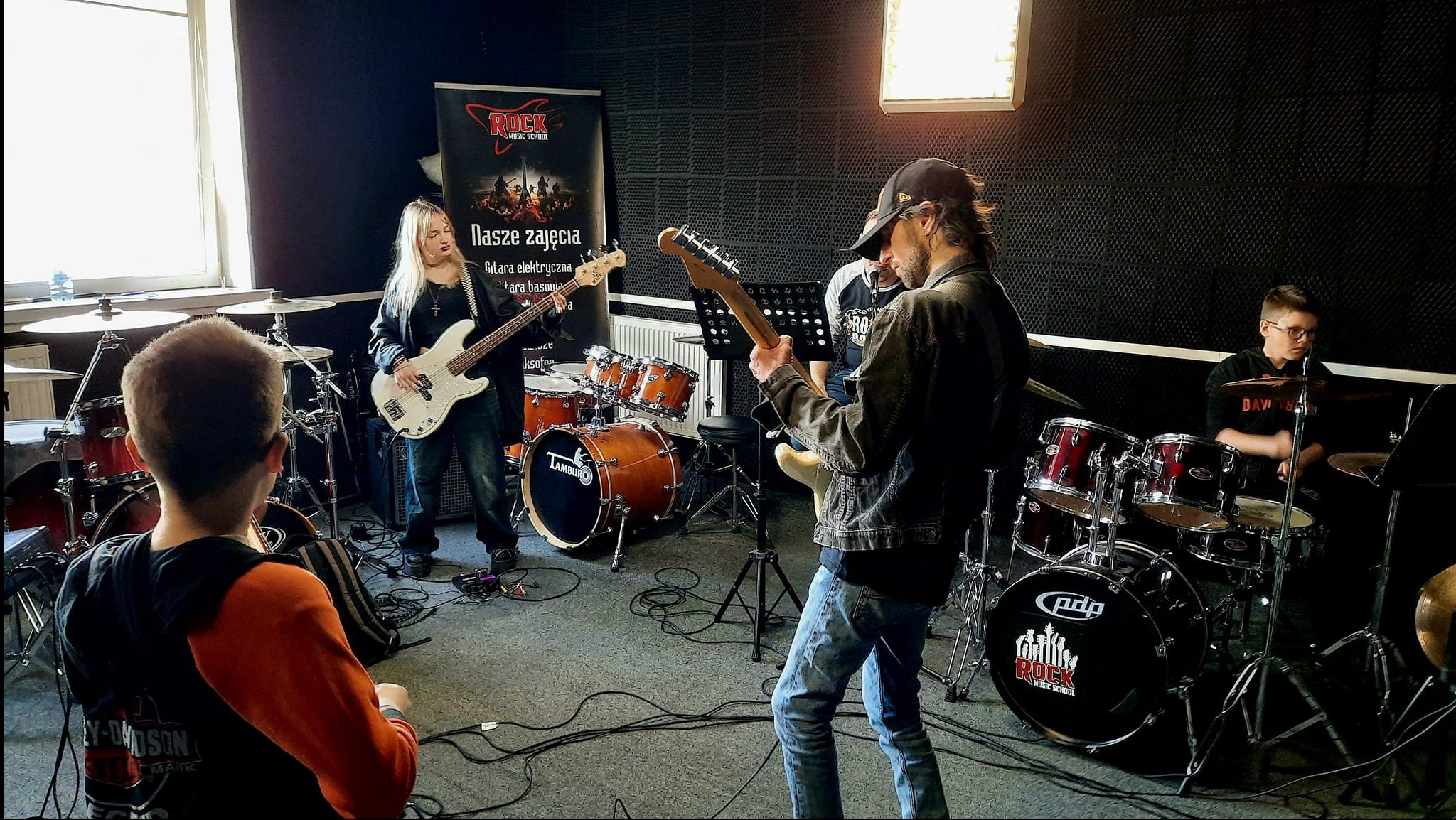 Zespół rockowy w trakcie próby w studiu: gitarzysta w jeansowej kurtce, basistka z białą gitarą basową, perkusista za zestawem perkusyjnym PDP Rock Music School, plakat Rock Music School w tle