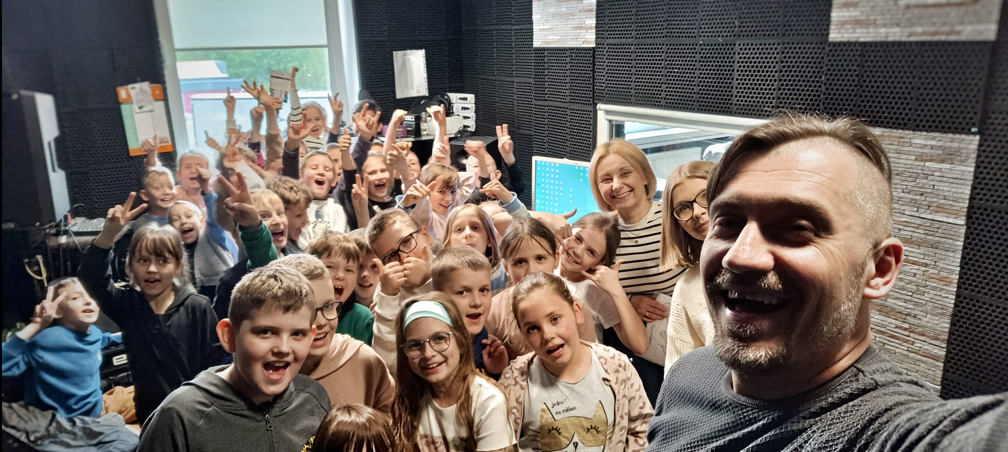 Radosna grupa dzieci i dorosłych w studiu nagrań, pozująca do selfie z uśmiechniętym mężczyzną, widoczne panele akustyczne na ścianach i sprzęt studyjny w tle.