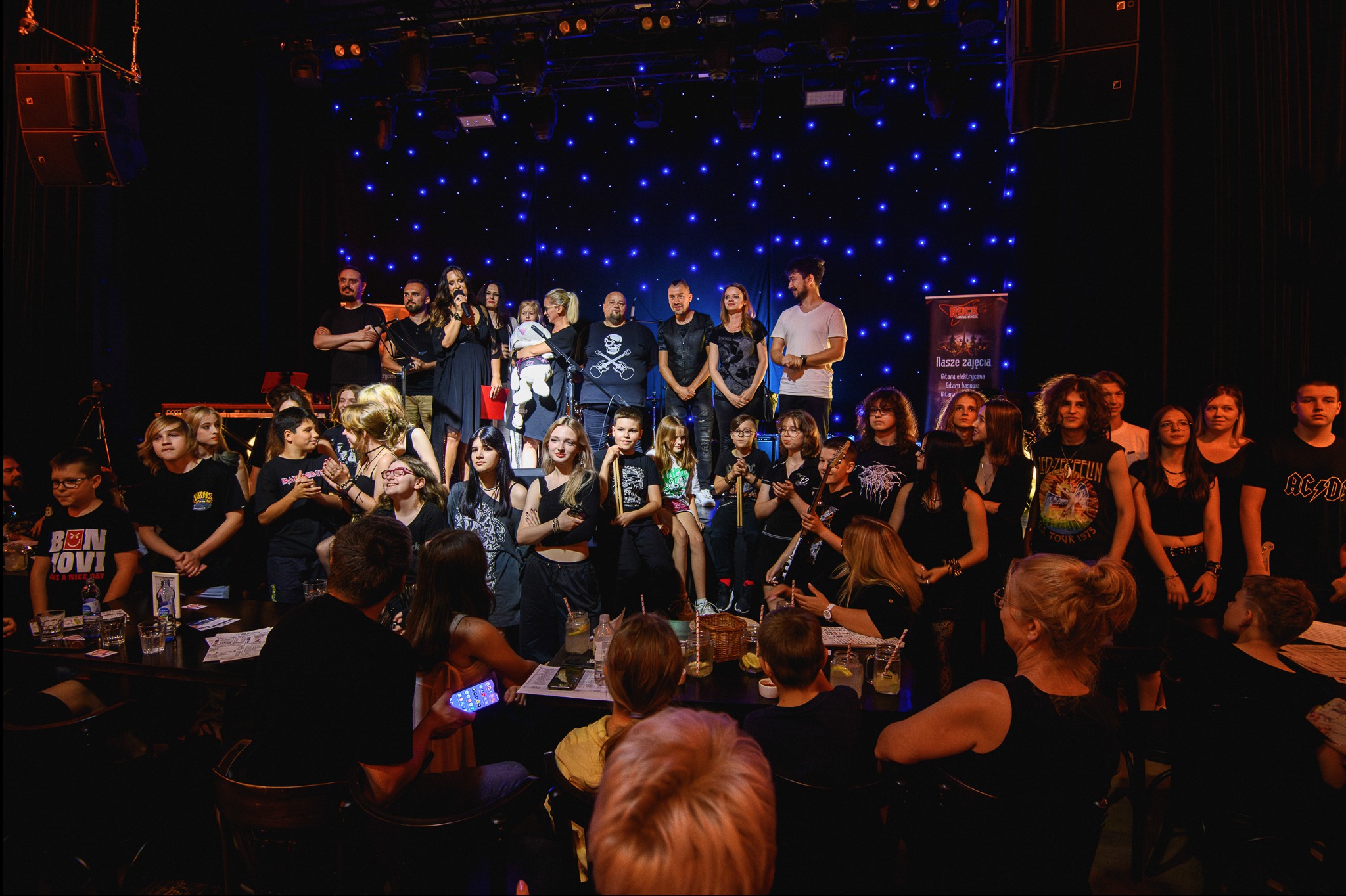 Grupowe zdjęcie uczestników i instruktorów szkoły rockowej na scenie z niebieskim oświetleniem, część osób siedzi przy stolikach z napojami.