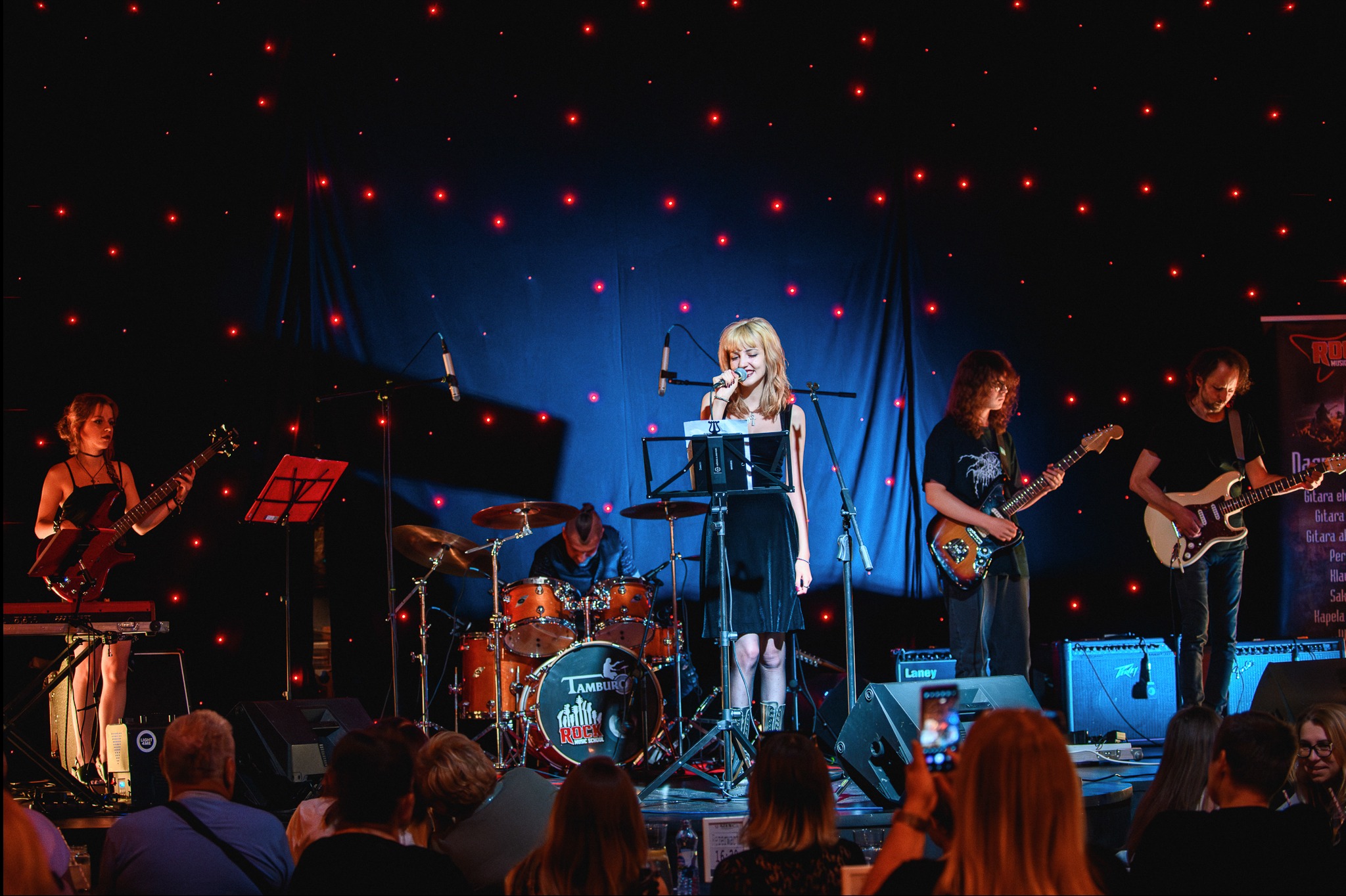 Zespół muzyczny na scenie podczas występu: wokalistka śpiewa do mikrofonu, gitarzyści grają na gitarach elektrycznych, perkusista za bębnami, a widzowie nagrywają występ telefonami.