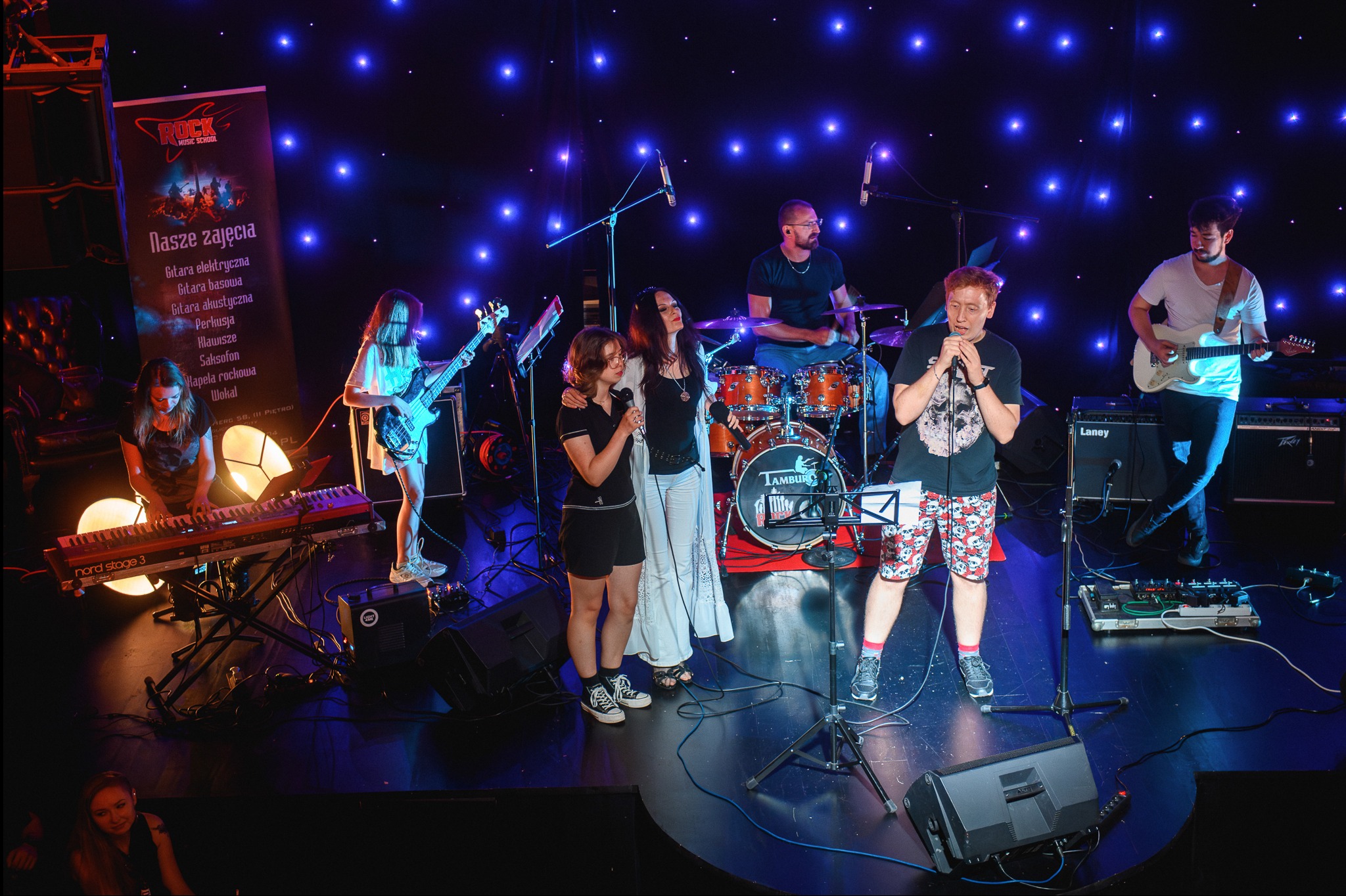 Zespół muzyczny na scenie podczas występu, wokalista w koszulce z czaszką i krótkich spodenkach w czaszki, dwie wokalistki, gitarzysta, basistka, klawiszowiec i perkusista, za nimi tło...