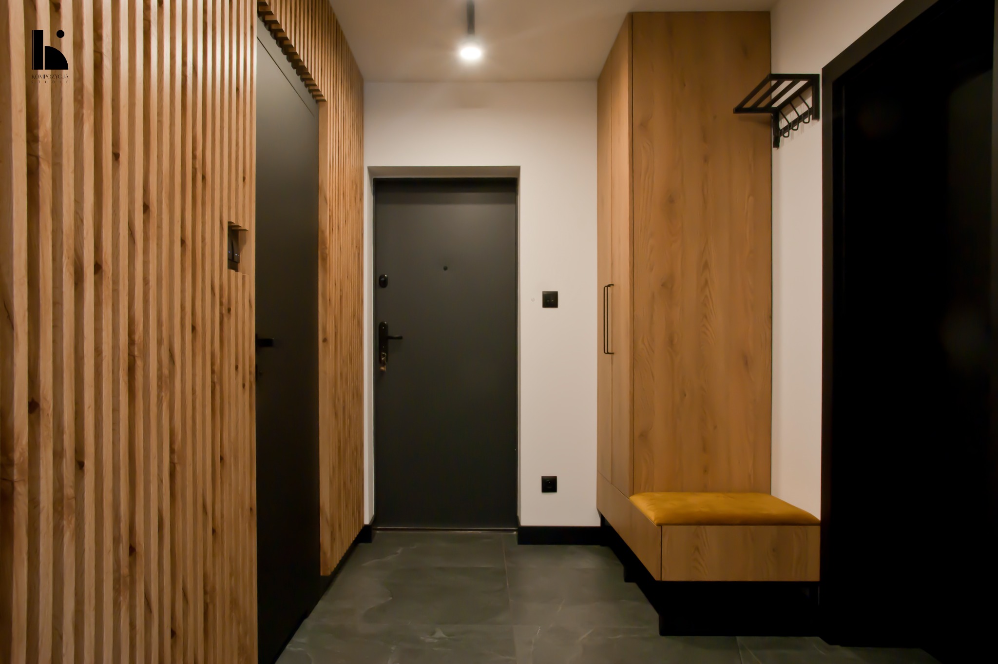 Nowoczesny przedpokój z drewnianymi lamelami na ścianie, szarą podłogą z płytek imitujących kamień i zabudowaną szafą z siedziskiem w kolorze musztardowym.